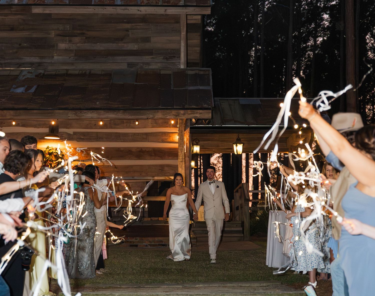Newlyweds exit wedding reception through sparkler and ribbon send-off at rustic barn venue at night.