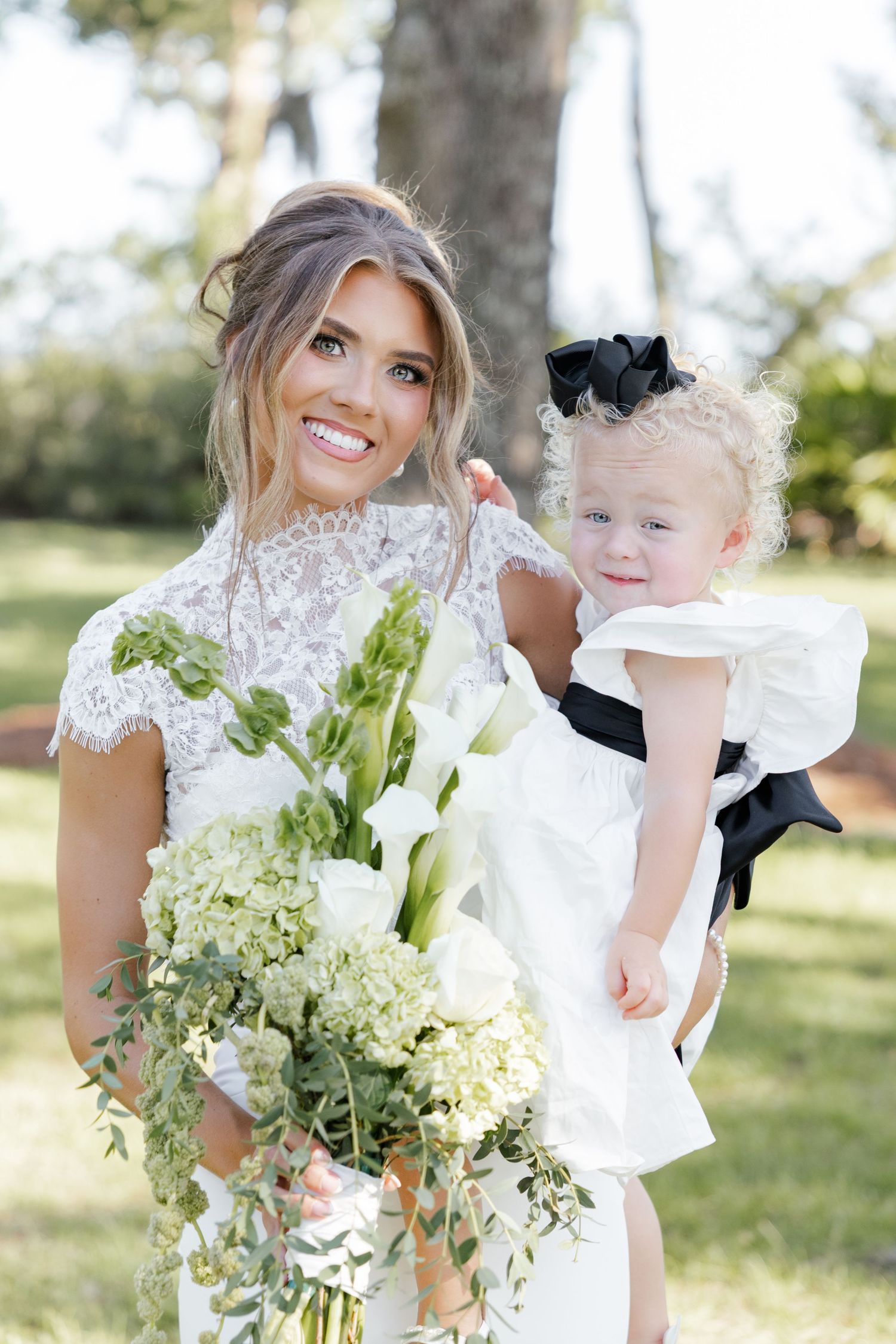 Smiling bride in lace wedding dress holds white floral bouquet and baby girl in white dress with black bow.