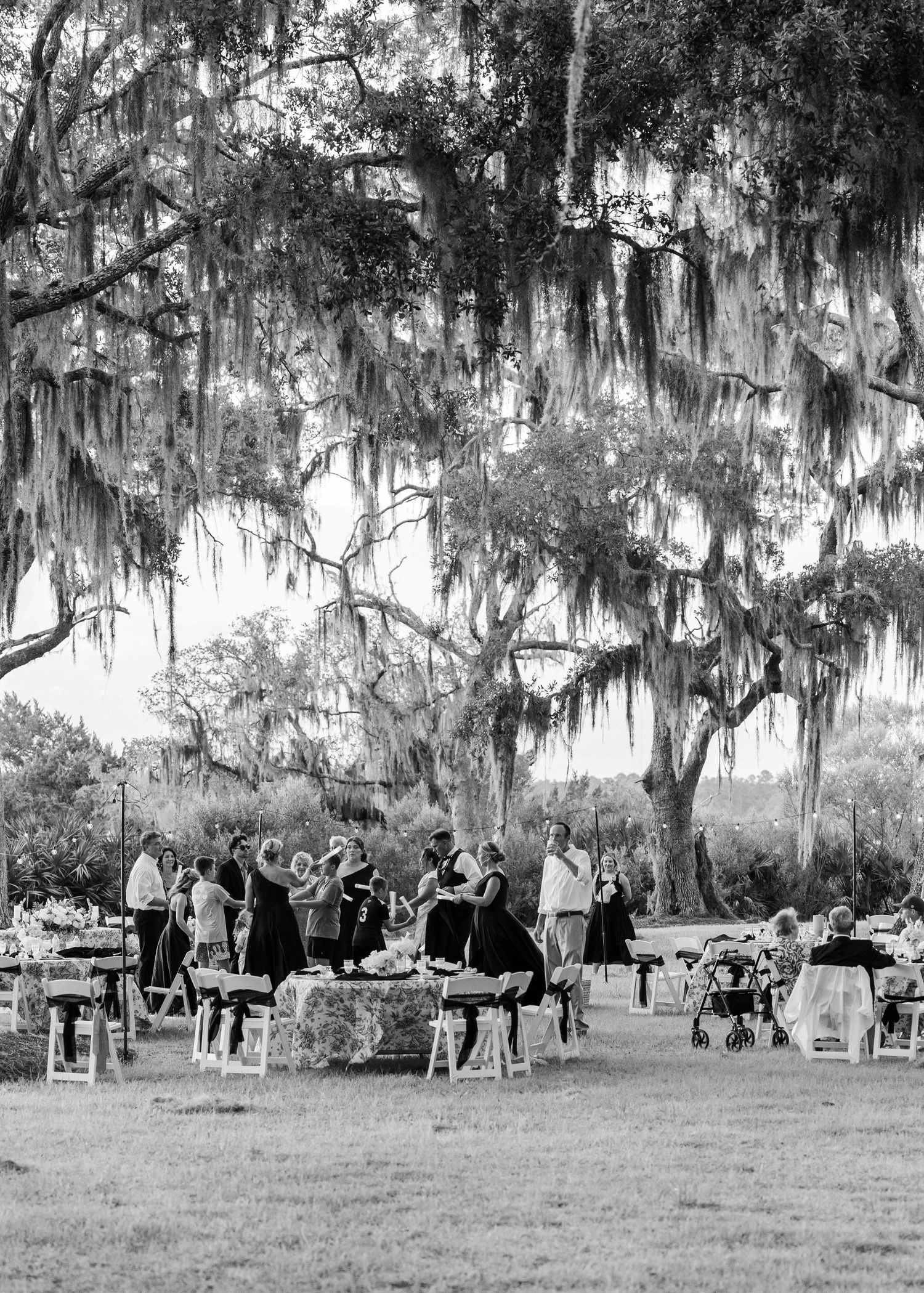 Black and white photo of wedding guests mingling outdoors under sprawling Spanish moss-draped oak trees.