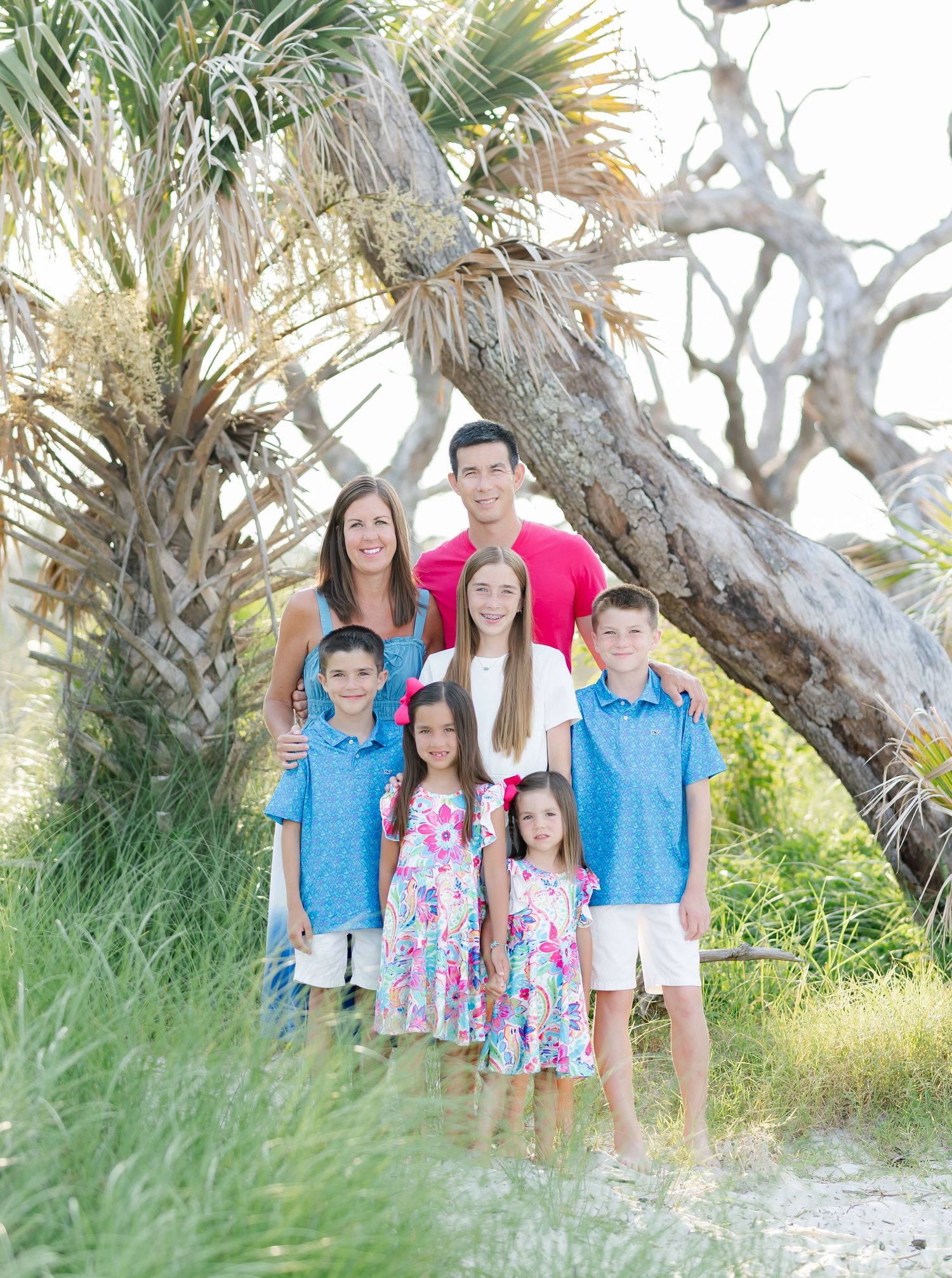 Family in coordinated blue and floral outfits poses together on a beach path with palm trees in the background.