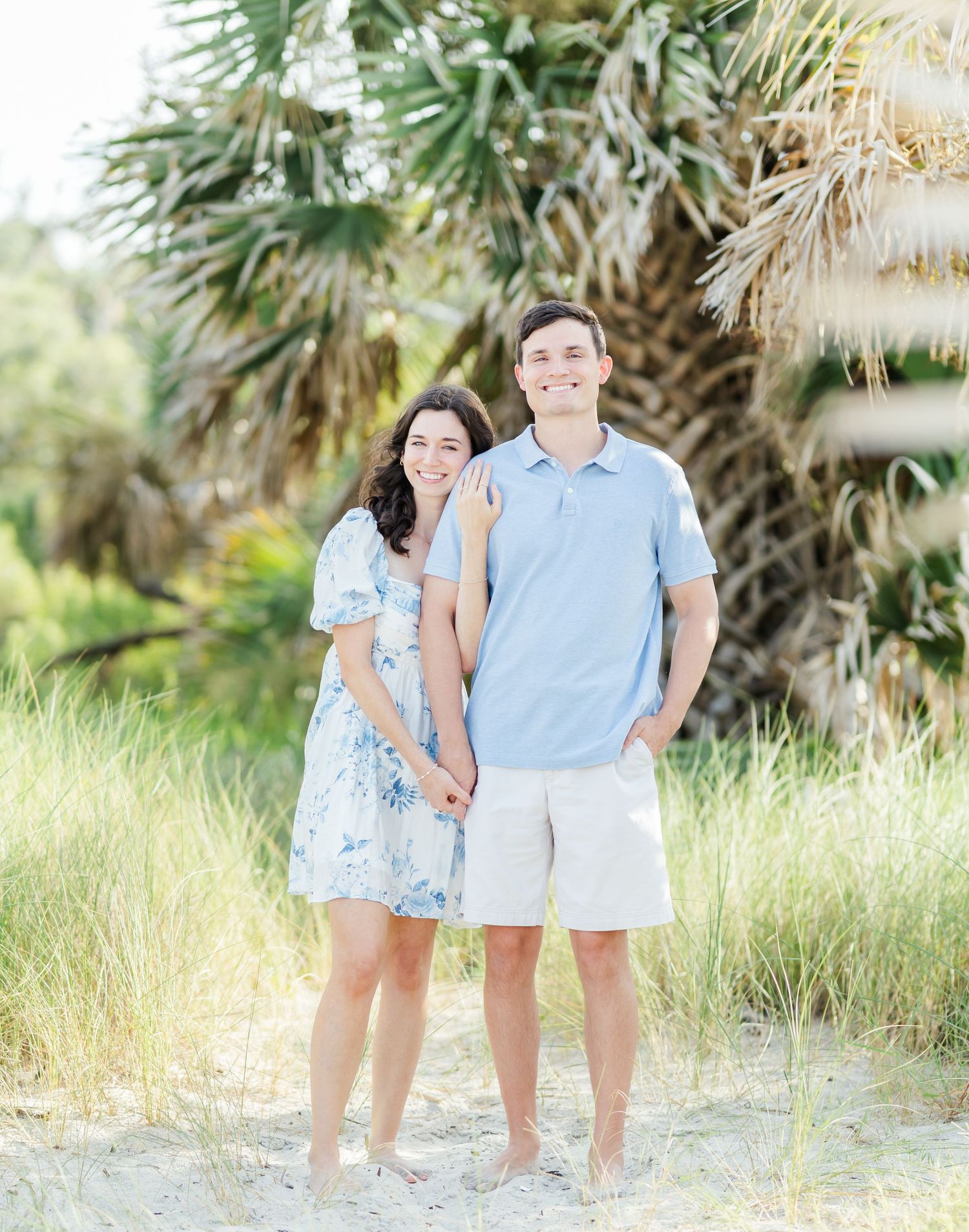 A couple in coordinating blue and white outfits pose together on a beach near palm trees.