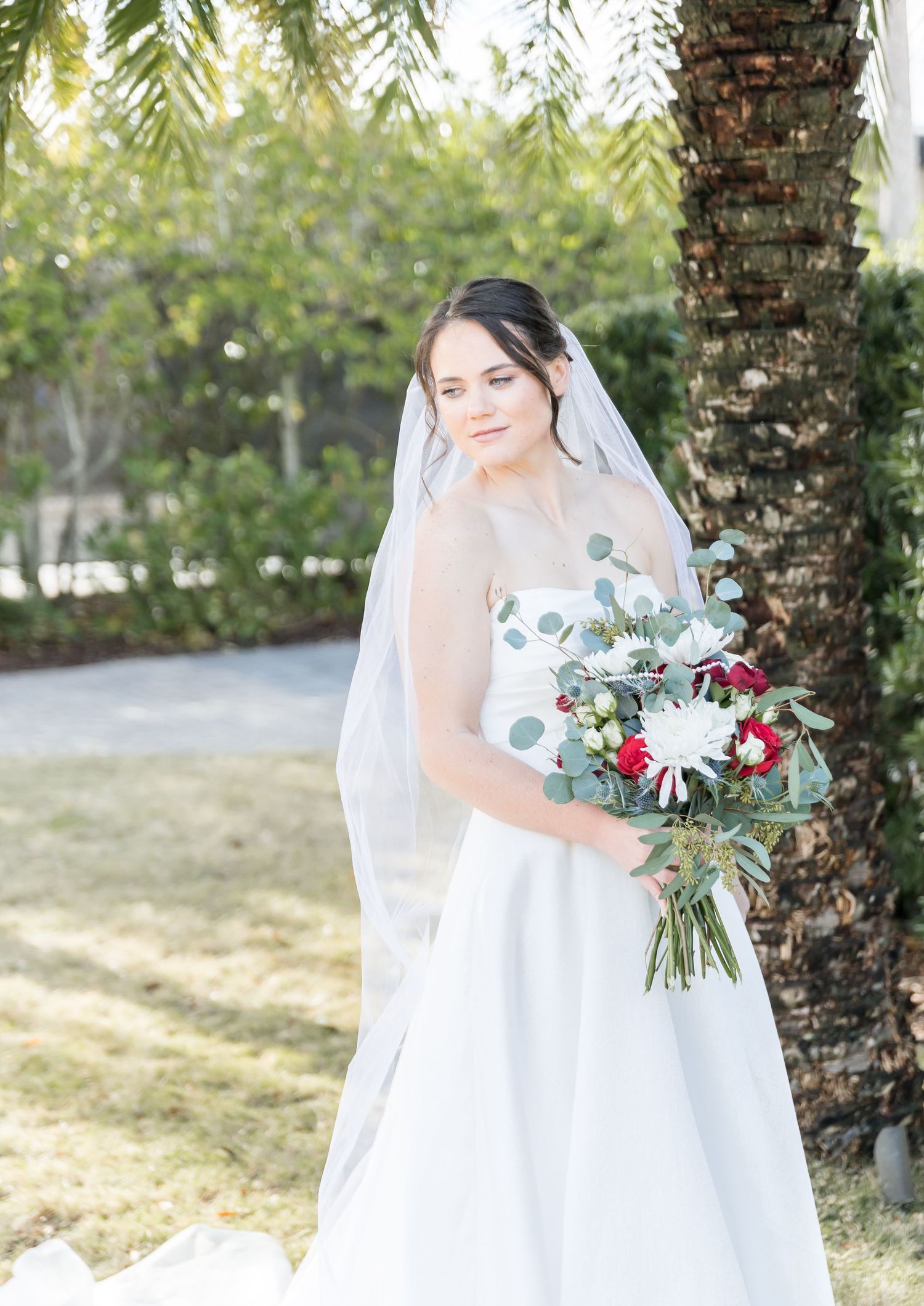 Bride in white wedding dress holds bouquet with red and white flowers outdoors.