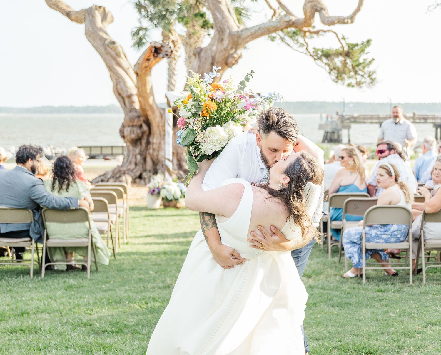Romantic outdoor wedding ceremony by the ocean with driftwood tree and floral arrangements on a sunny day.