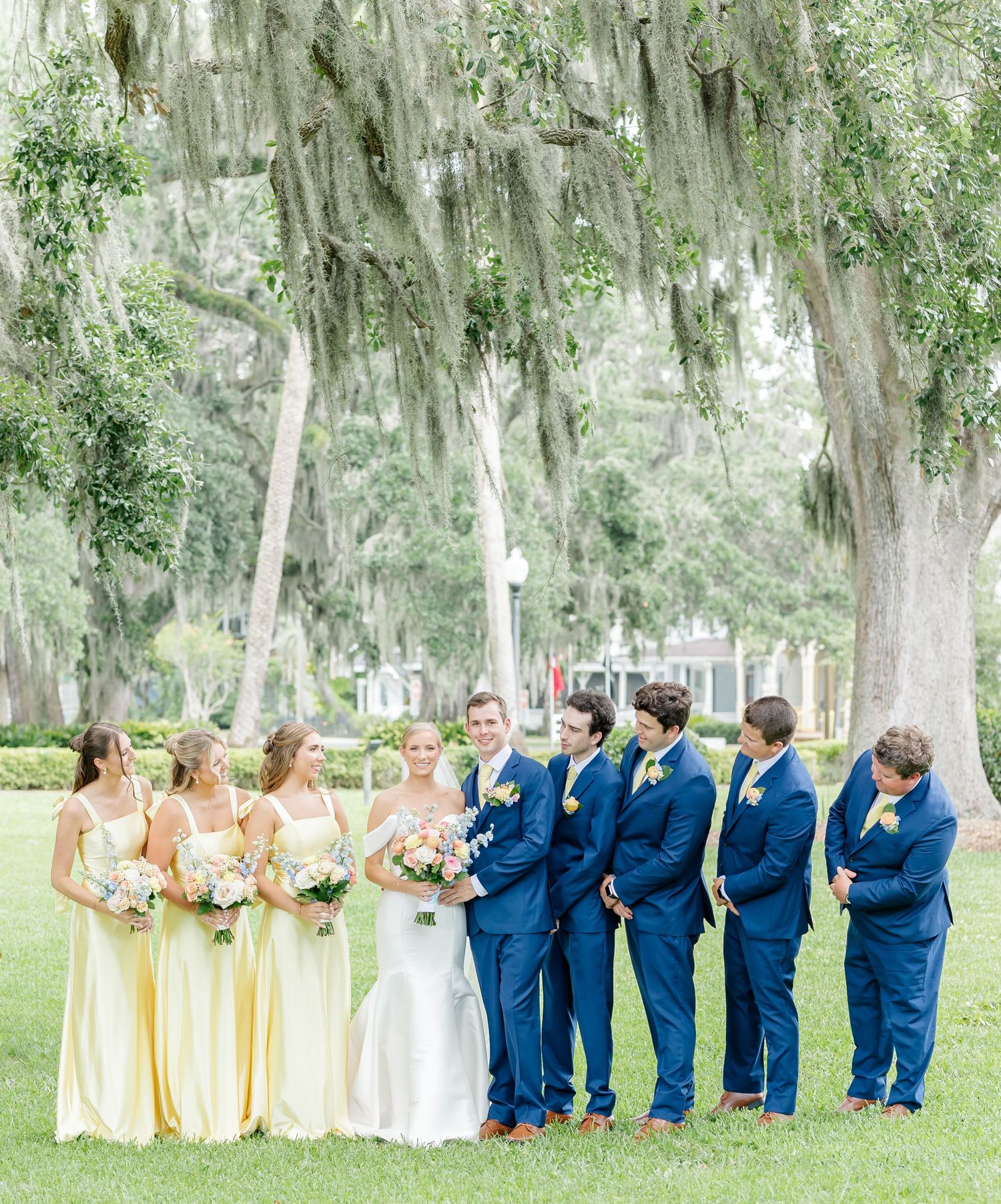 Bridal party in navy suits and pale yellow dresses lined up under moss-covered oak trees.