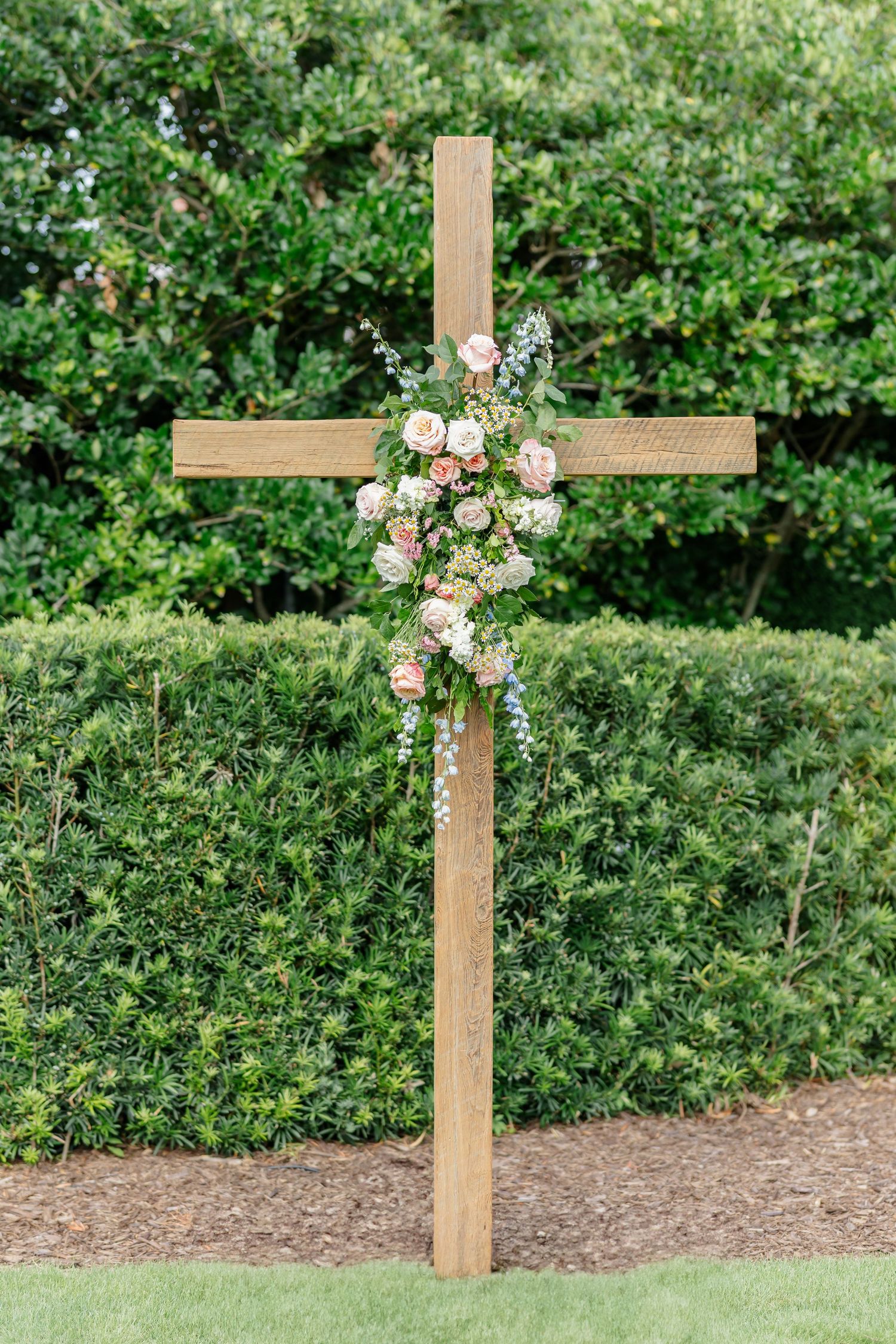 A wooden cross decorated with pink roses and flowers stands against lush green garden foliage.