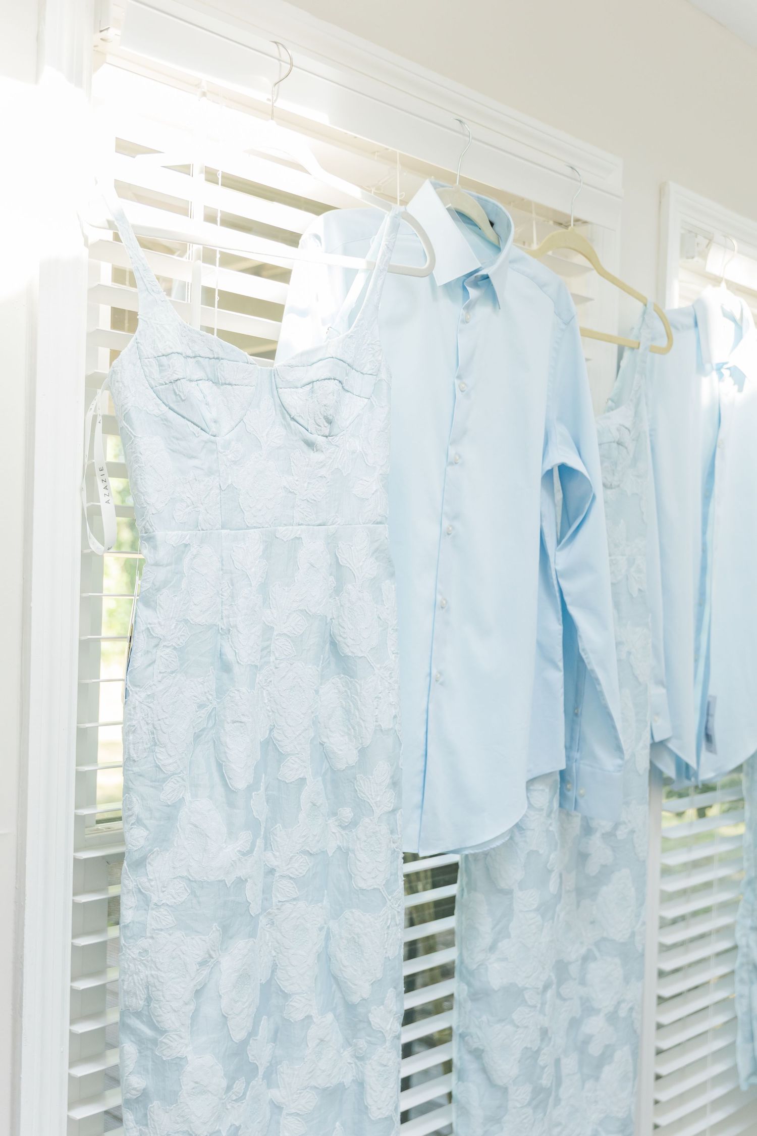 Row of delicate light blue lace dresses hanging on white hangers by a sunny window.