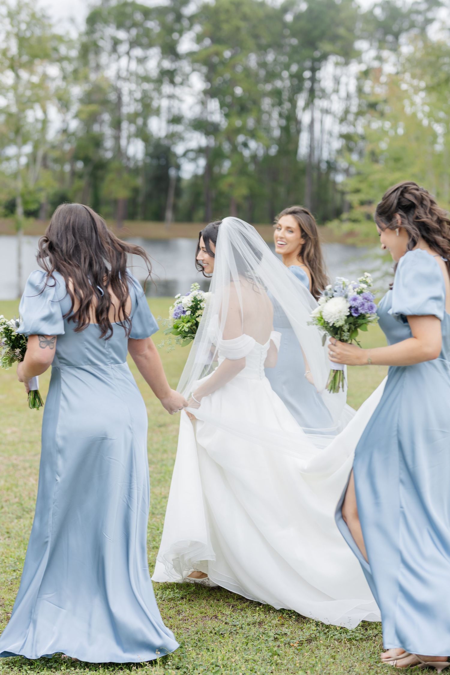 Wedding party in light blue dresses and white gown pose outdoors by a lake while holding floral bouquets.