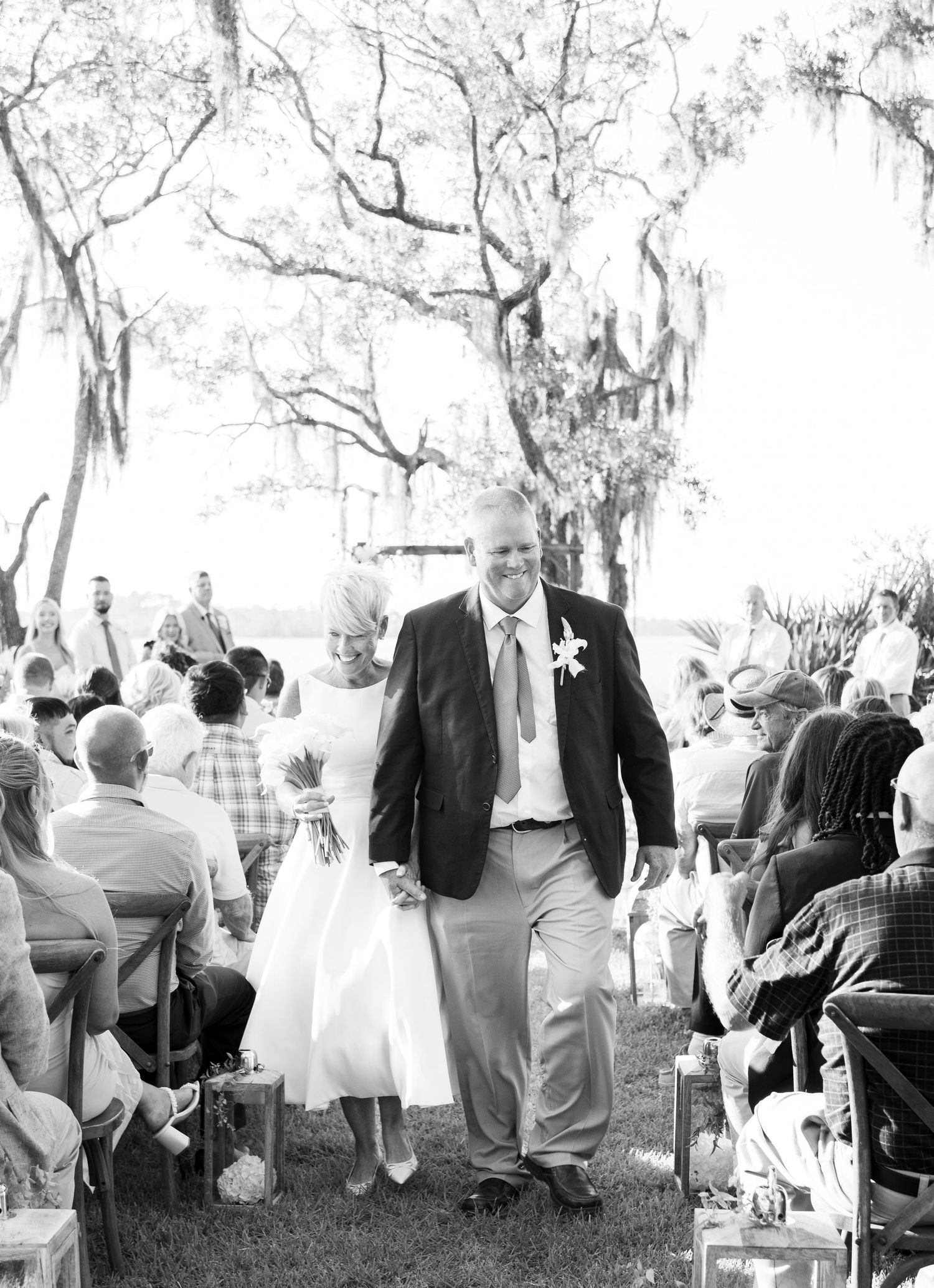 Black and white photo of wedding guests gathered for an outdoor ceremony under moss-covered trees.