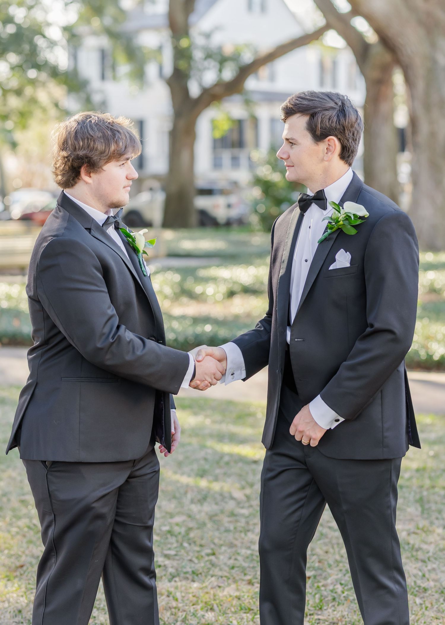 Groomsmen in black tuxedos share handshakes during outdoor wedding photos.