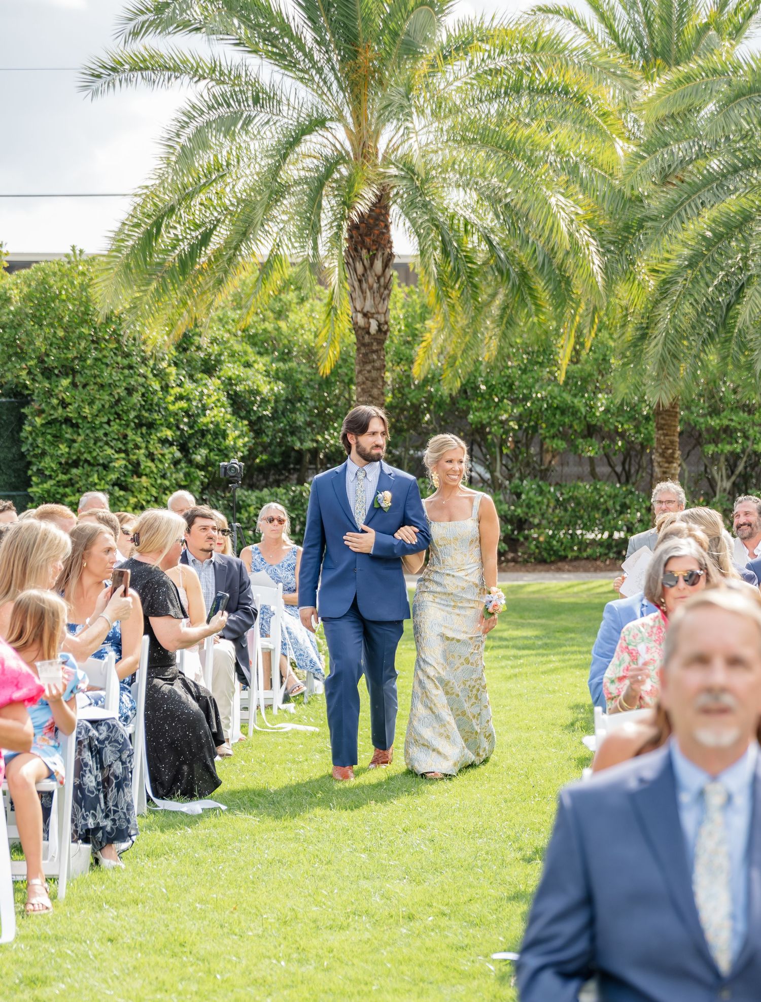 Outdoor wedding ceremony under palm trees with guests seated on white chairs in a tropical garden setting.