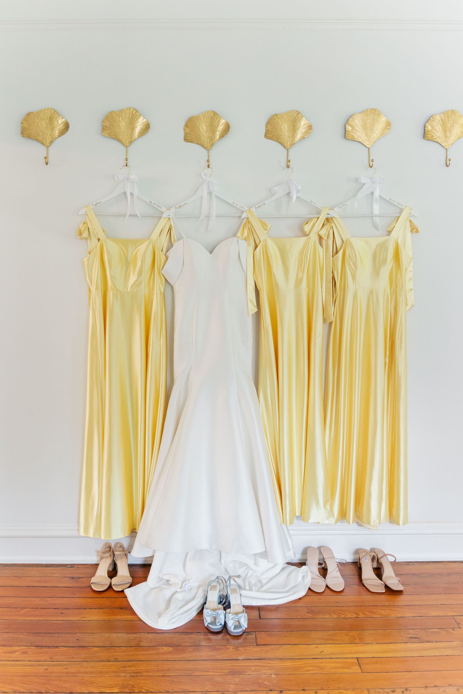 Yellow bridesmaid dresses and a white wedding gown hanging on display with matching shoes beneath on hardwood floor.
