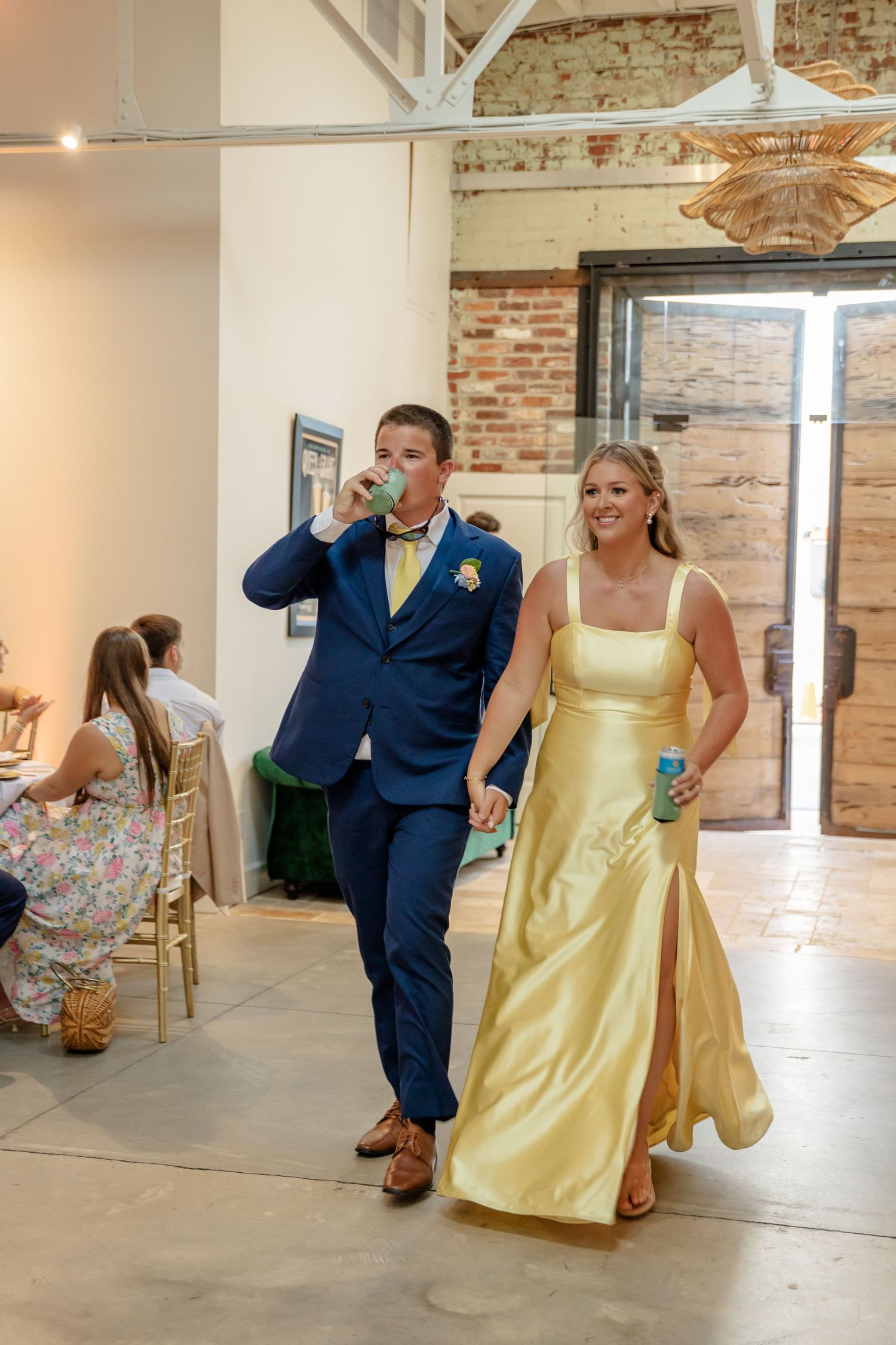 Wedding couple in navy and yellow attire playfully dance together in a rustic brick-walled venue.