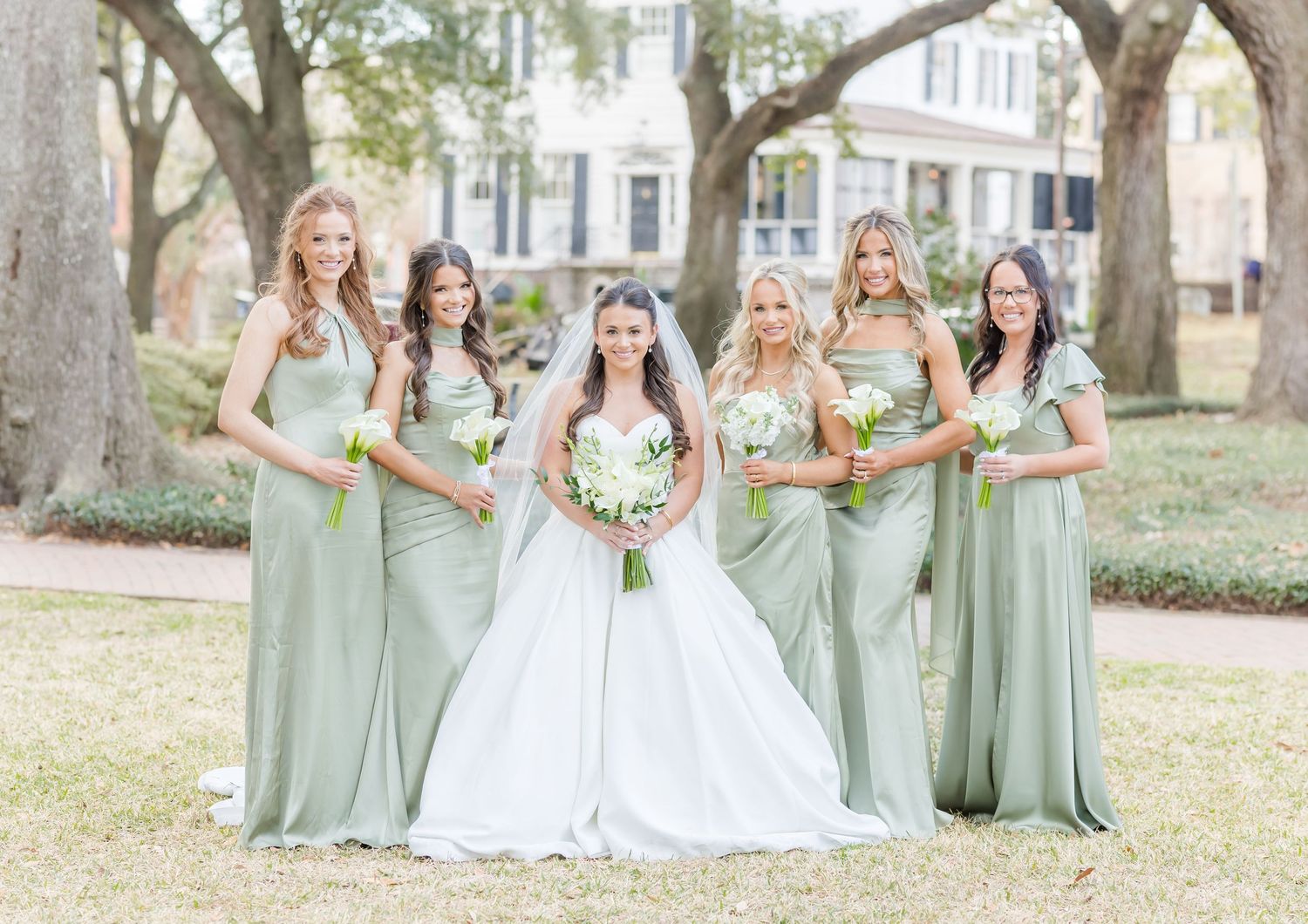 Wedding party in sage green bridesmaid dresses poses together outdoors under oak trees.