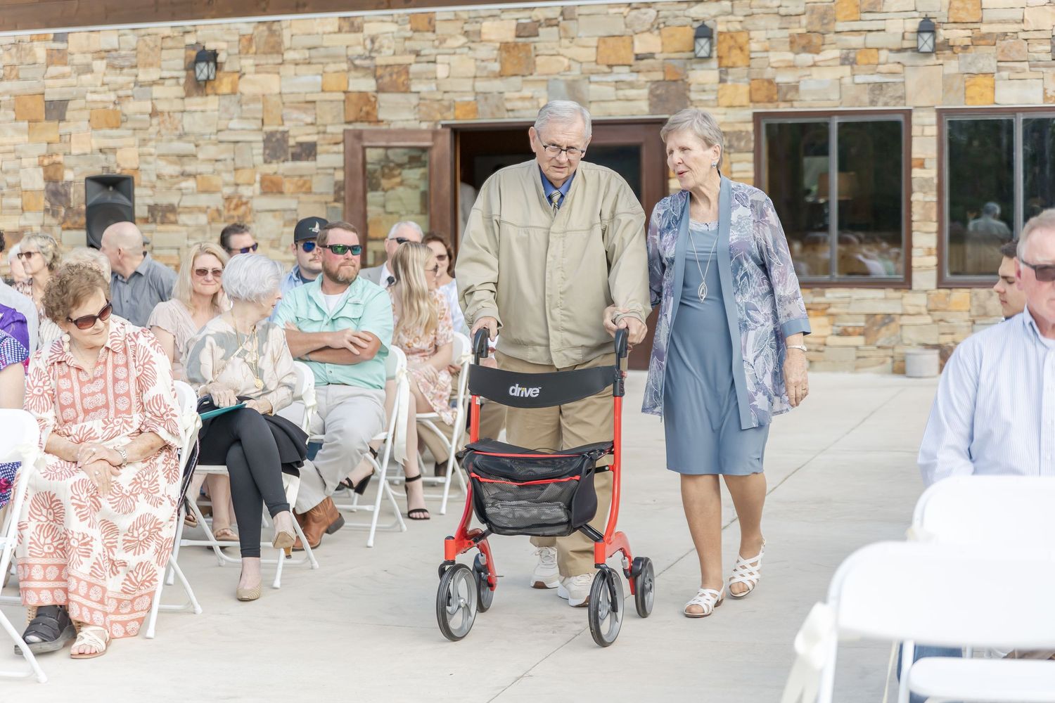 Wedding guests seated in white chairs outside stone building during ceremony.