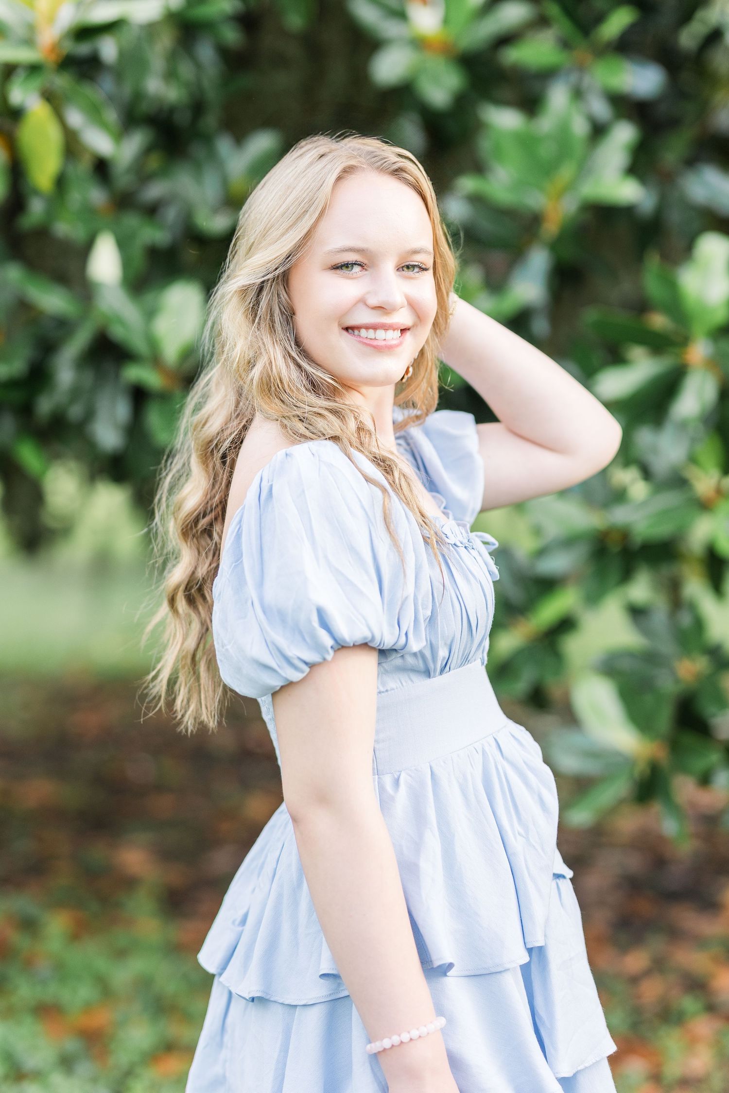 A blonde woman in a light blue ruffled dress poses outdoors against a green garden background.