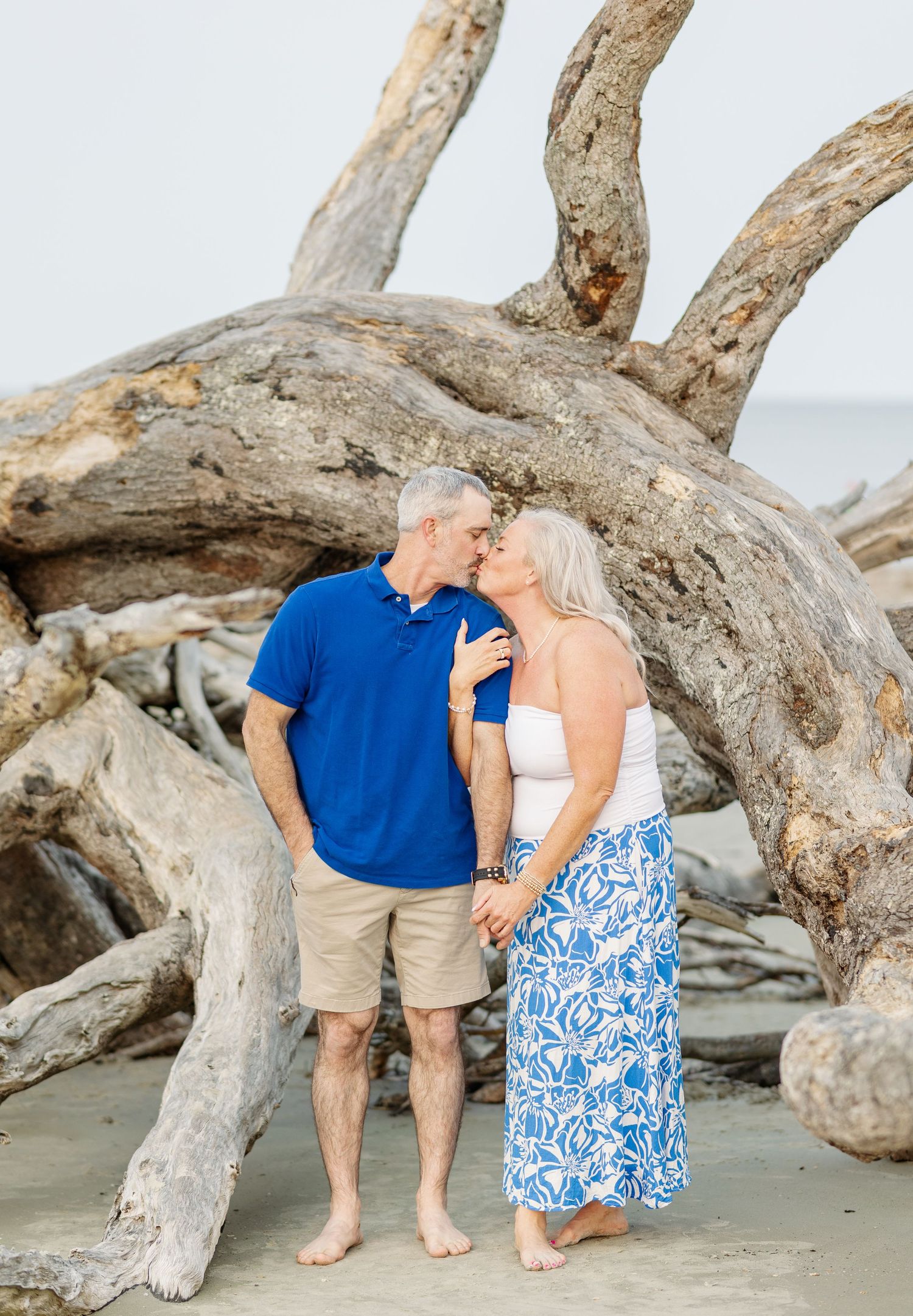 A couple shares an intimate moment on a beach with driftwood and weathered logs in the background.
