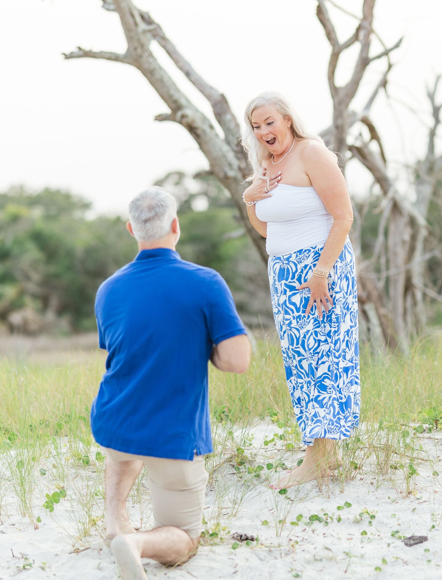 A surprise proposal moment captured on a sandy beach with driftwood trees in the background.