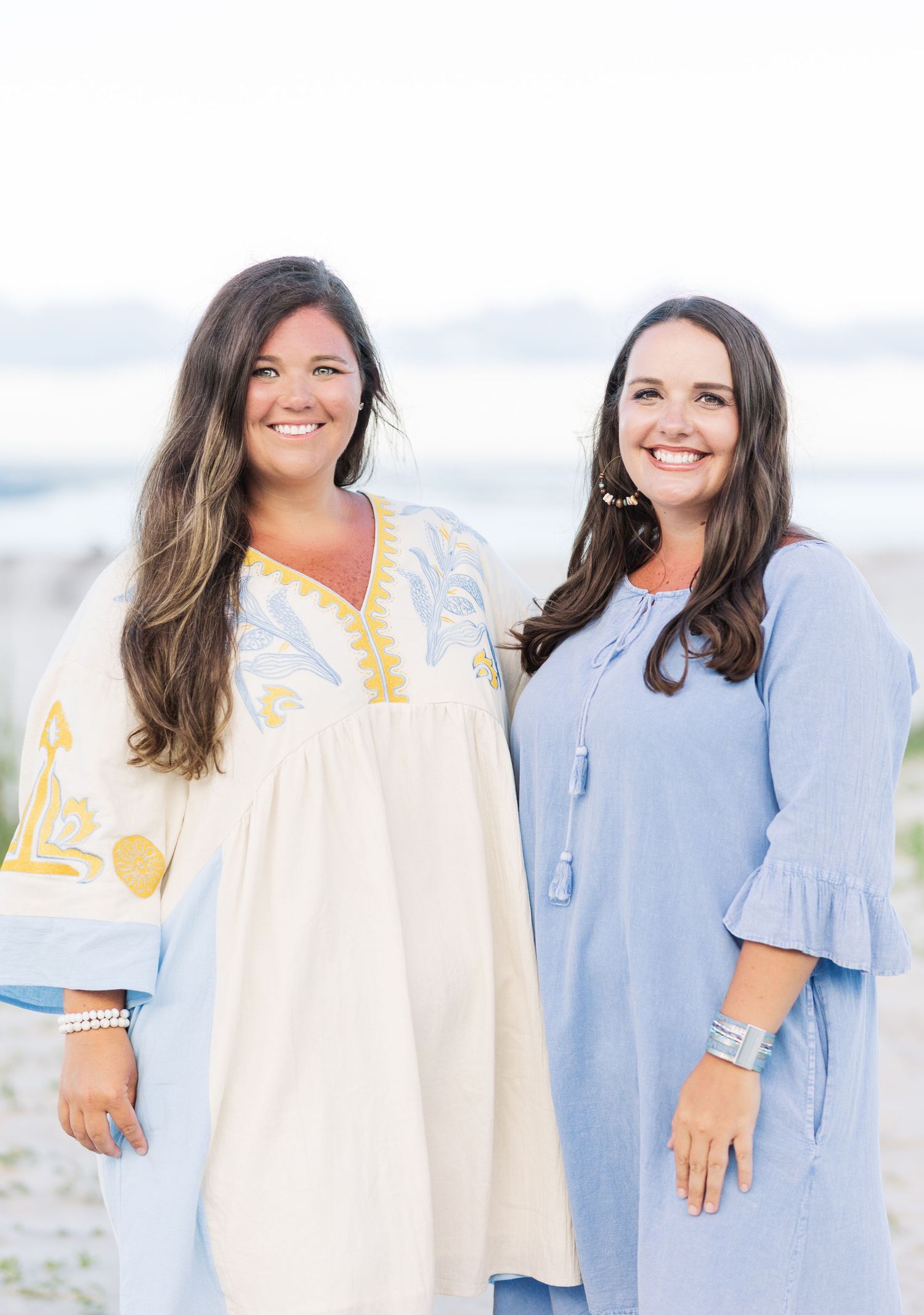 Four women in coordinated blue and white summer dresses pose together on beach during sunset.