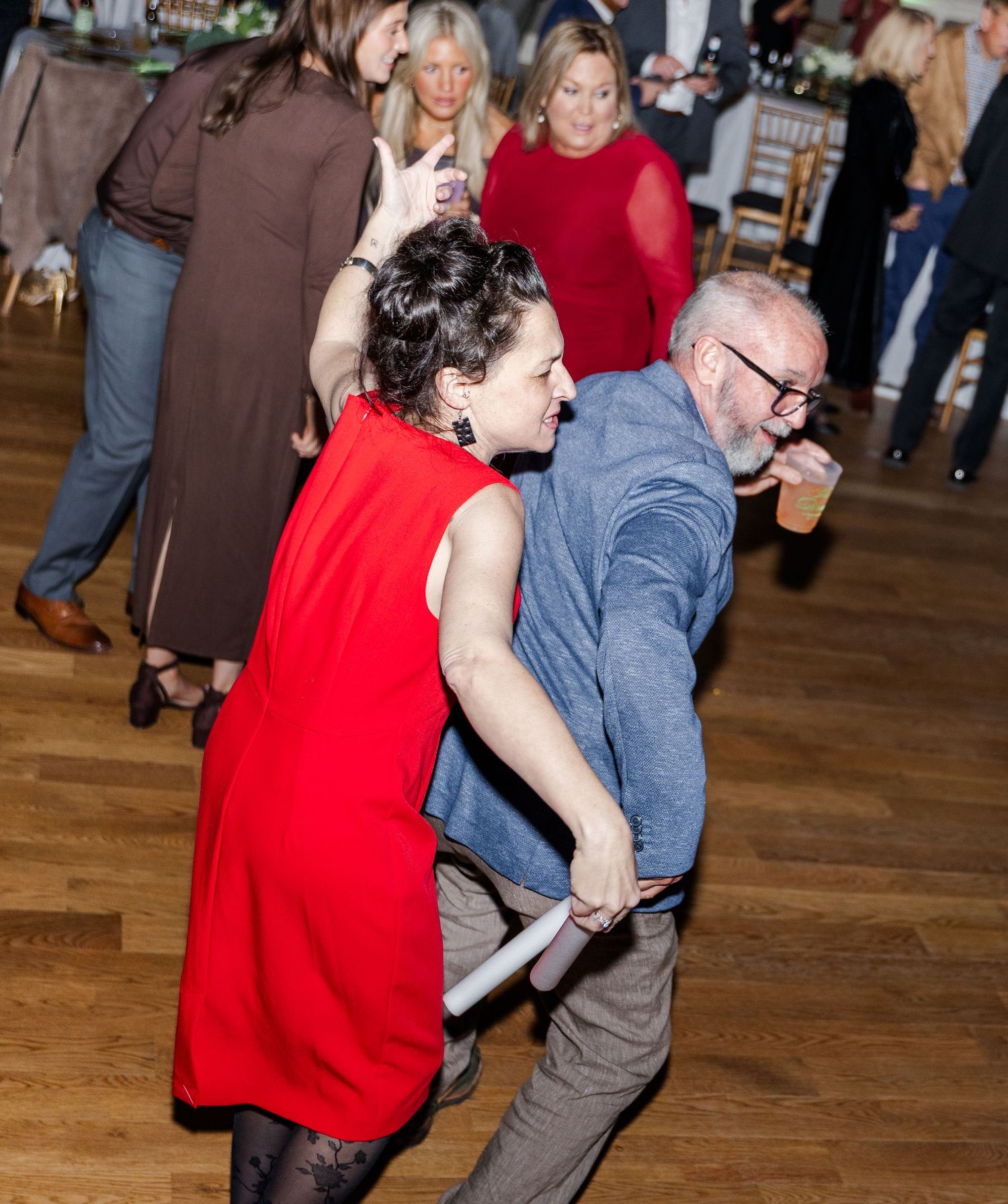 Guests dance energetically at a wedding reception on a hardwood floor in a festive venue.