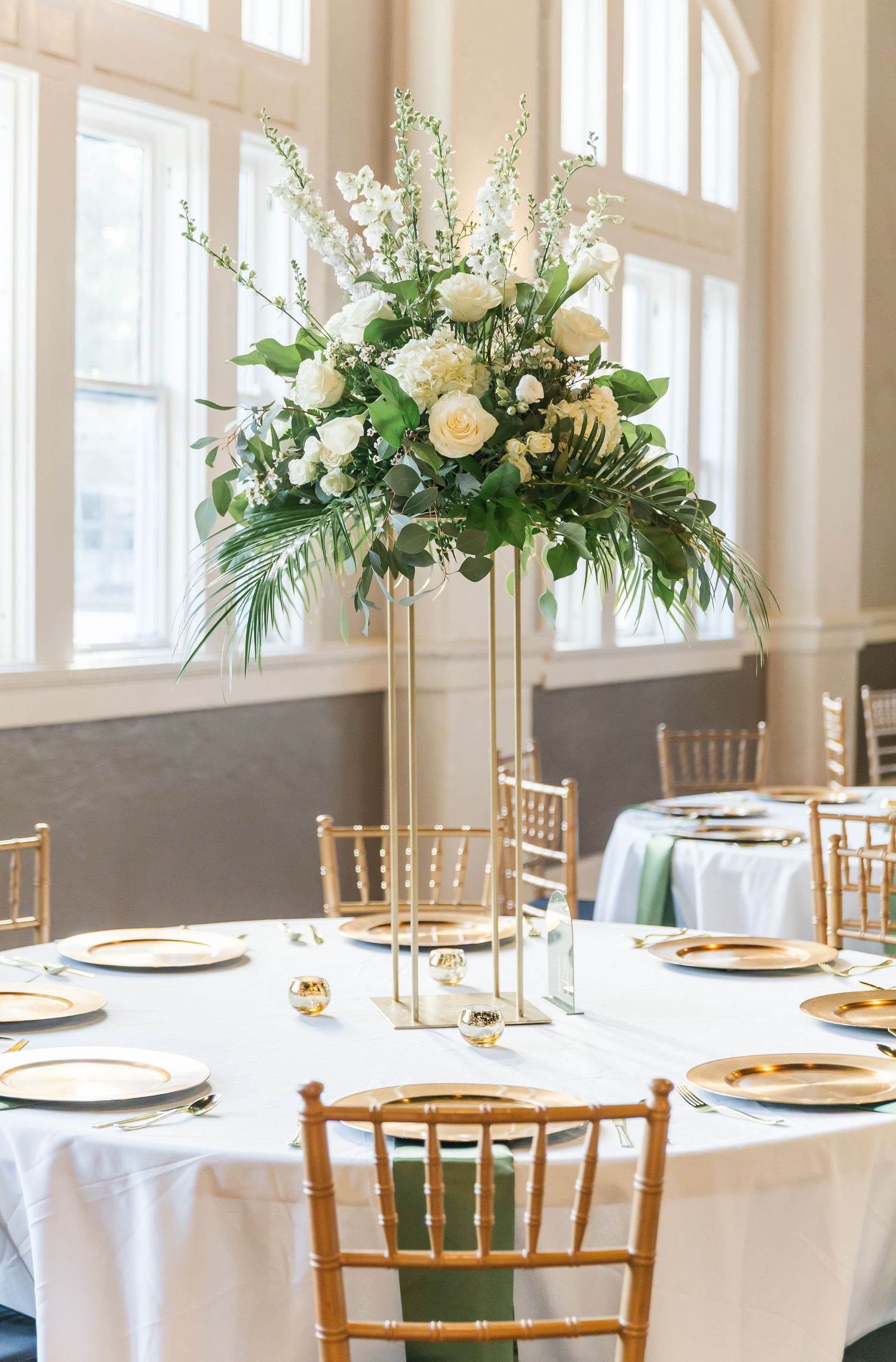 Tall gold pillar centerpiece with white roses, delphinium and tropical greenery at sunlit wedding reception table.