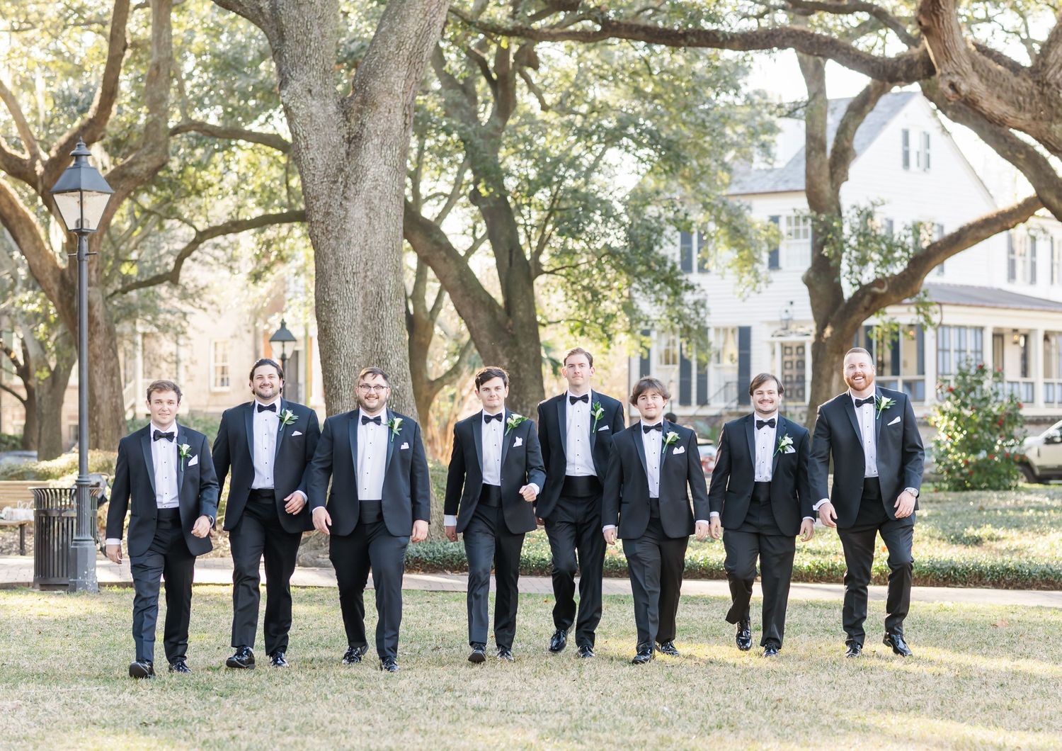 Groom and groomsmen in black tuxedos walk together outdoors under oak trees at a wedding.