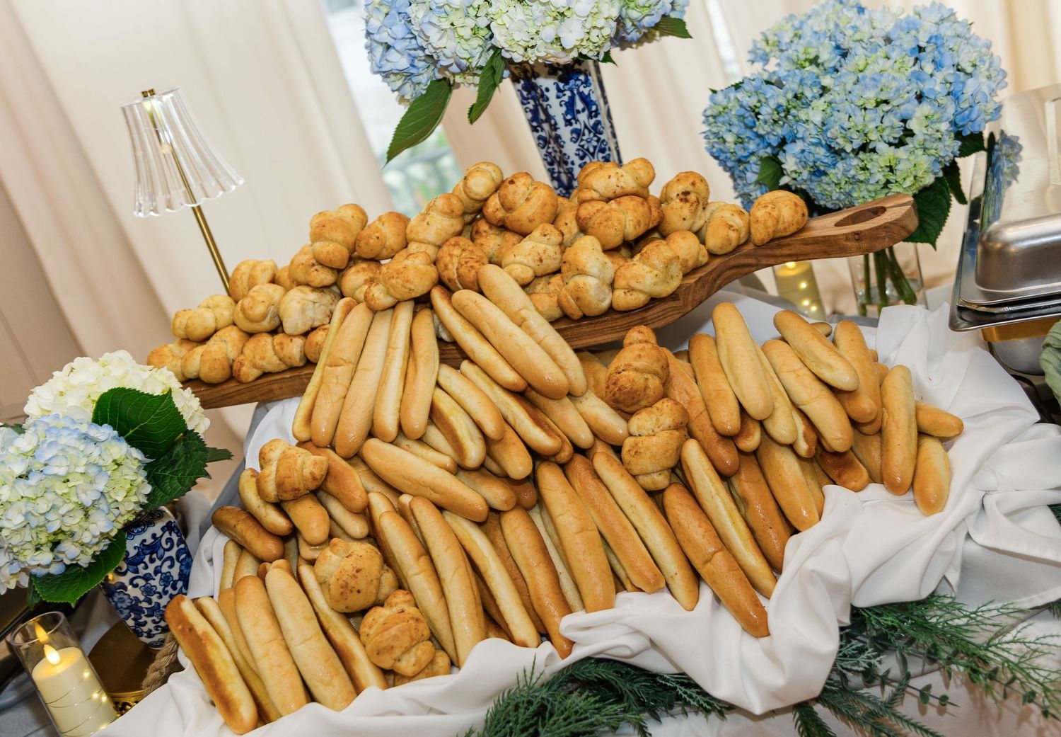 Elegant buffet display of breadsticks and dinner rolls arranged on a white draped table with blue hydrangeas.