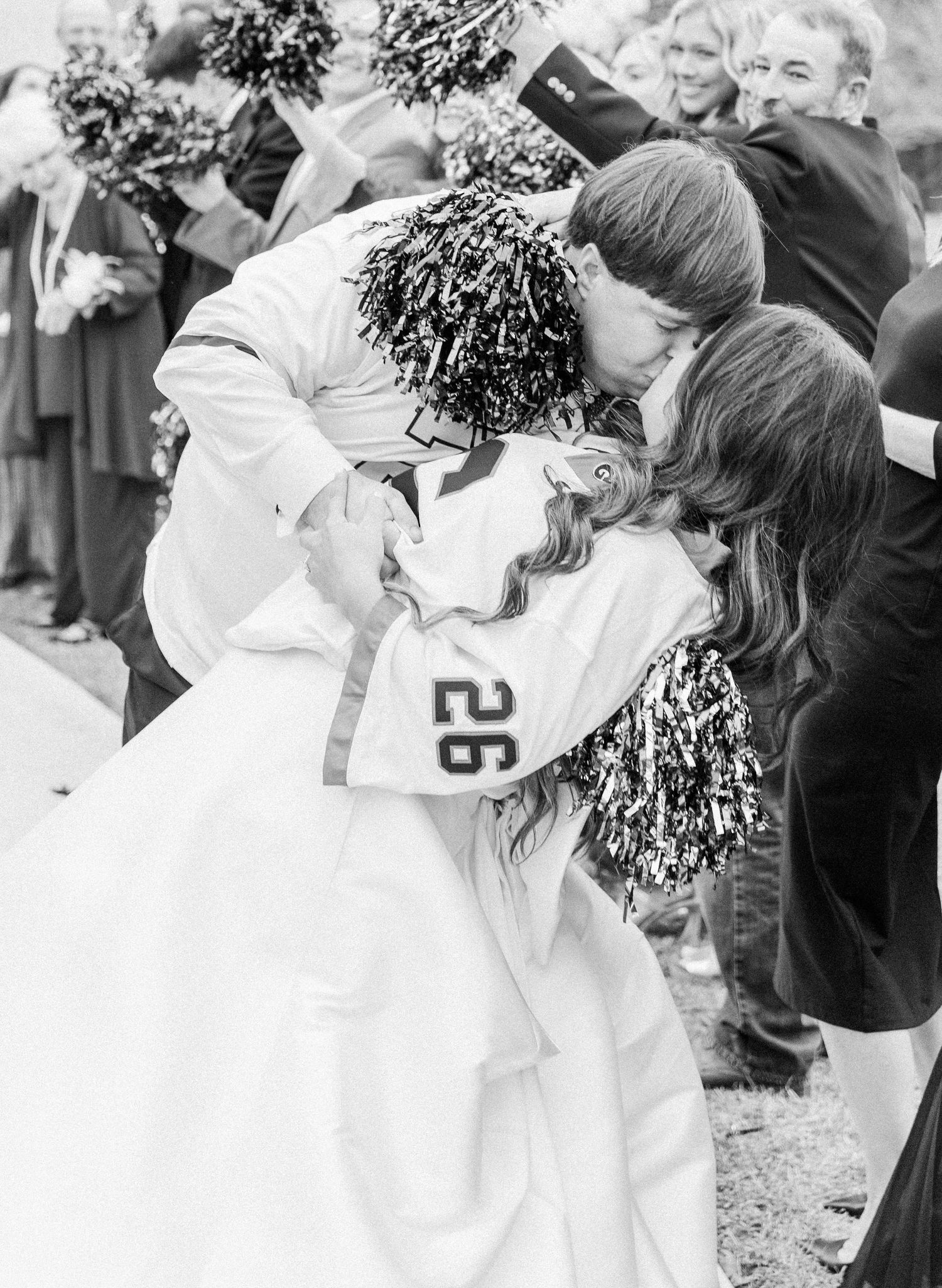 Black and white photo of bride and groom sharing a kiss during festive football-themed wedding exit.