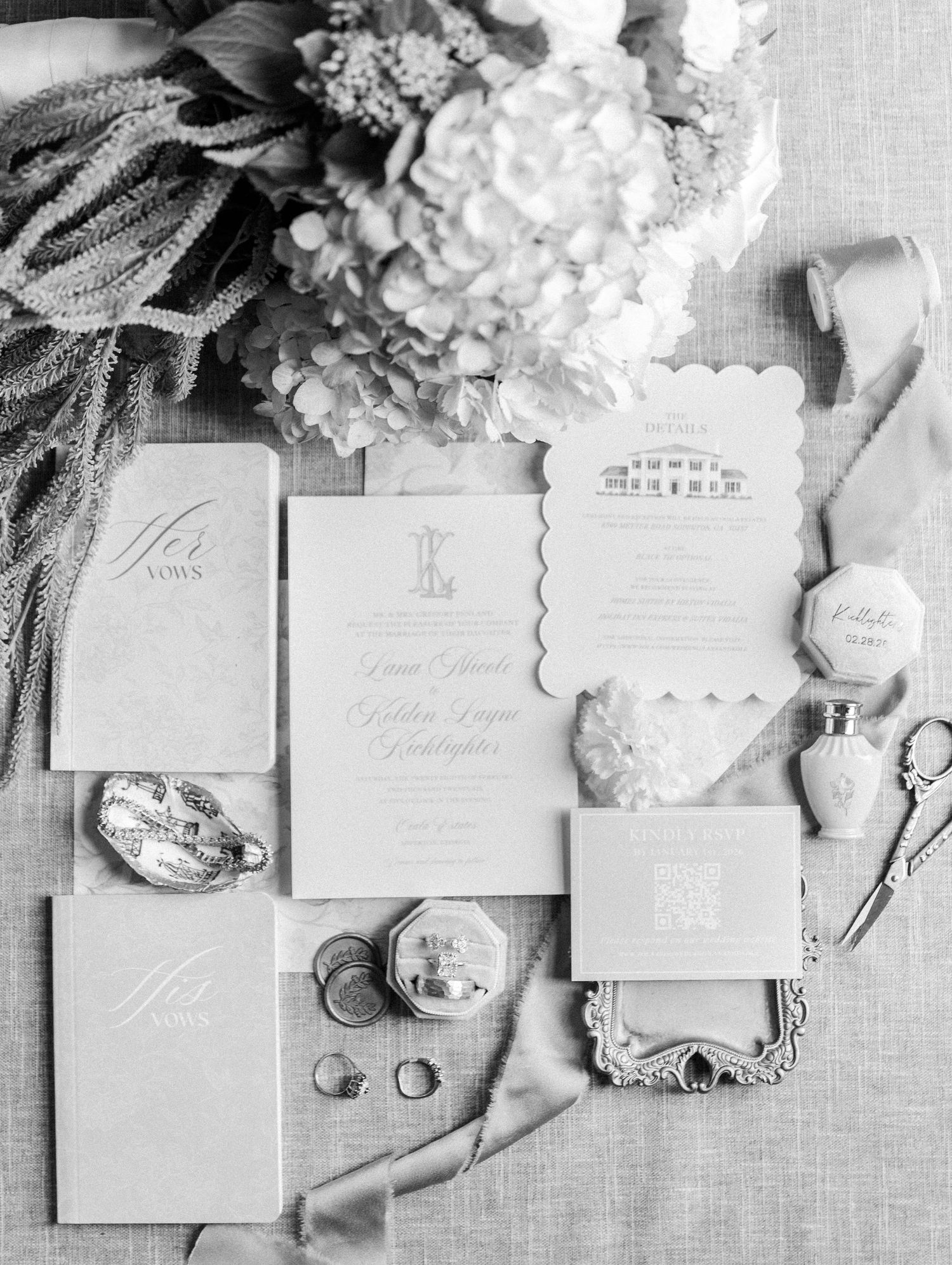 Black and white flat lay of elegant wedding invitation suite with rings, ribbon, and floral accents.