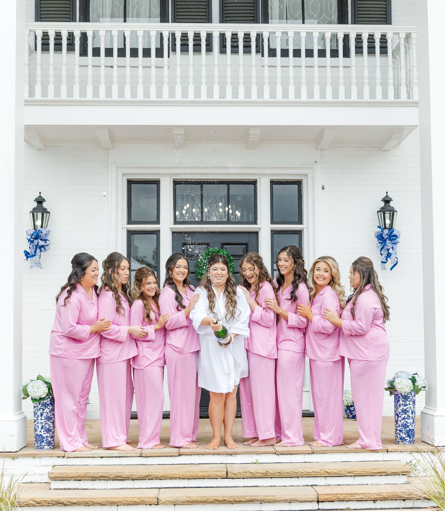 Bride in white robe holding champagne bottle flanked by bridesmaids in pink pajamas outside a classic white Southern home.