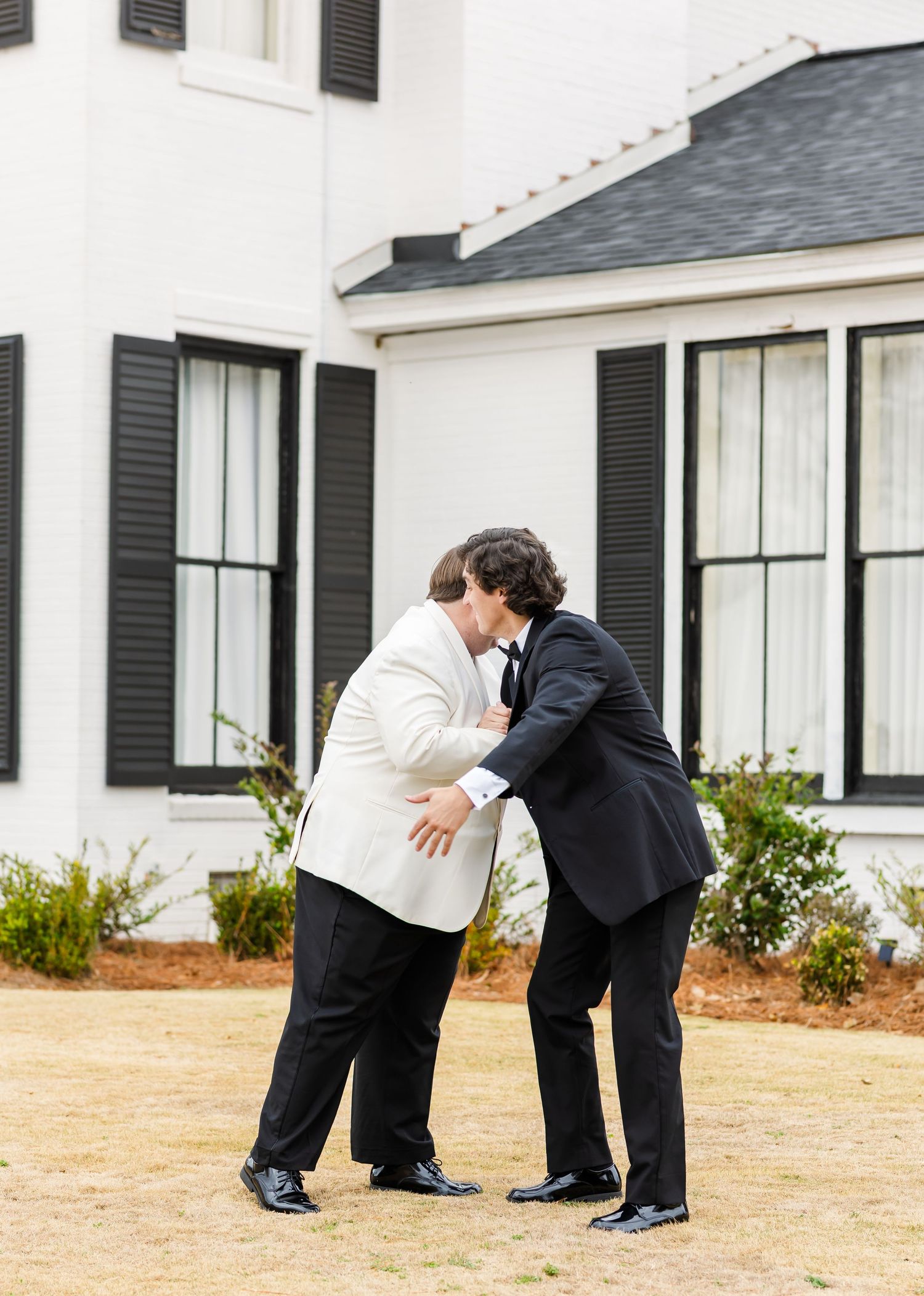 Groom in white jacket shares a playful moment with groomsman in black tuxedo outside white venue.