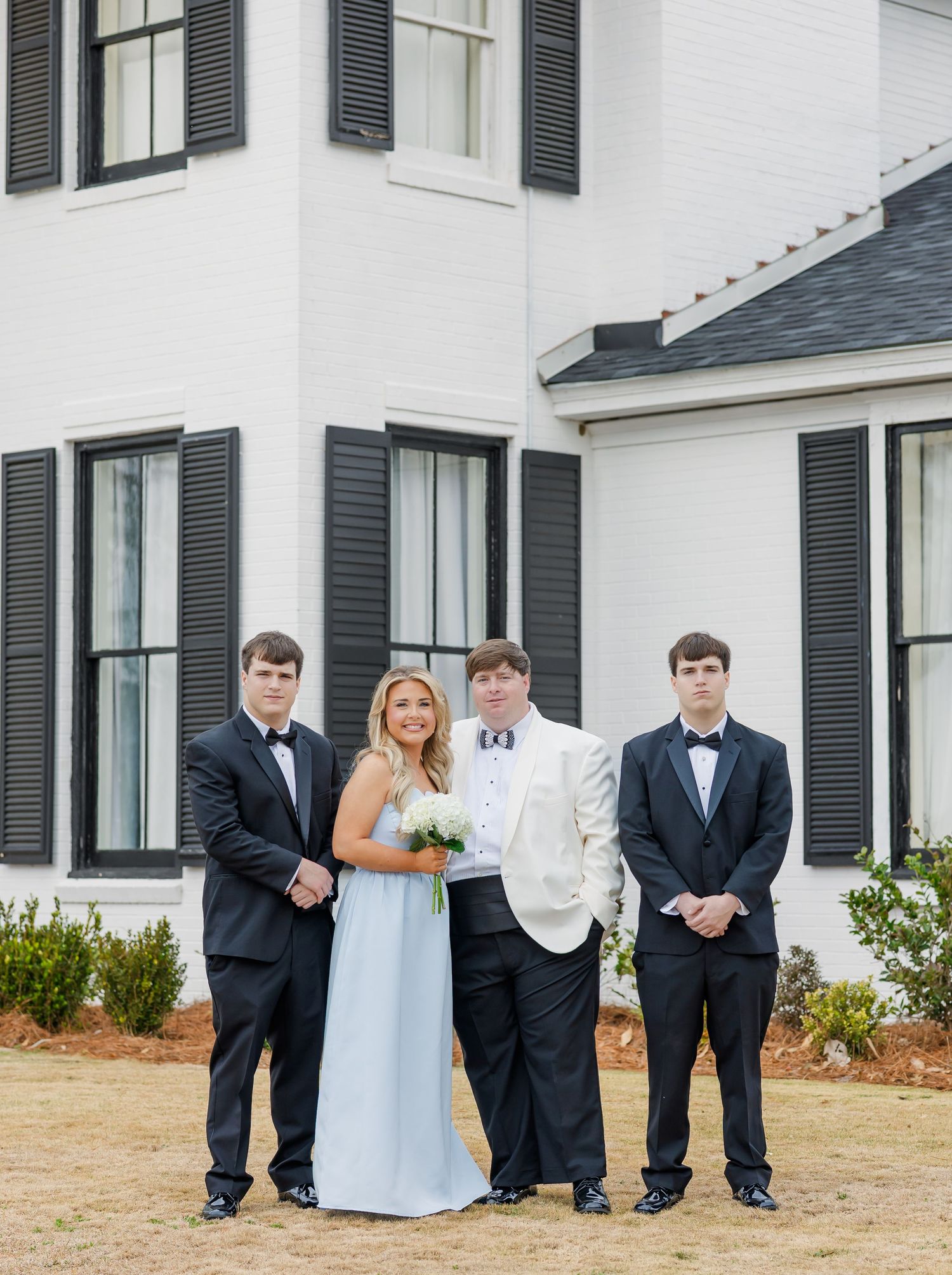 Groom in white tuxedo with bride in light blue dress flanked by two groomsmen in black tuxedos.