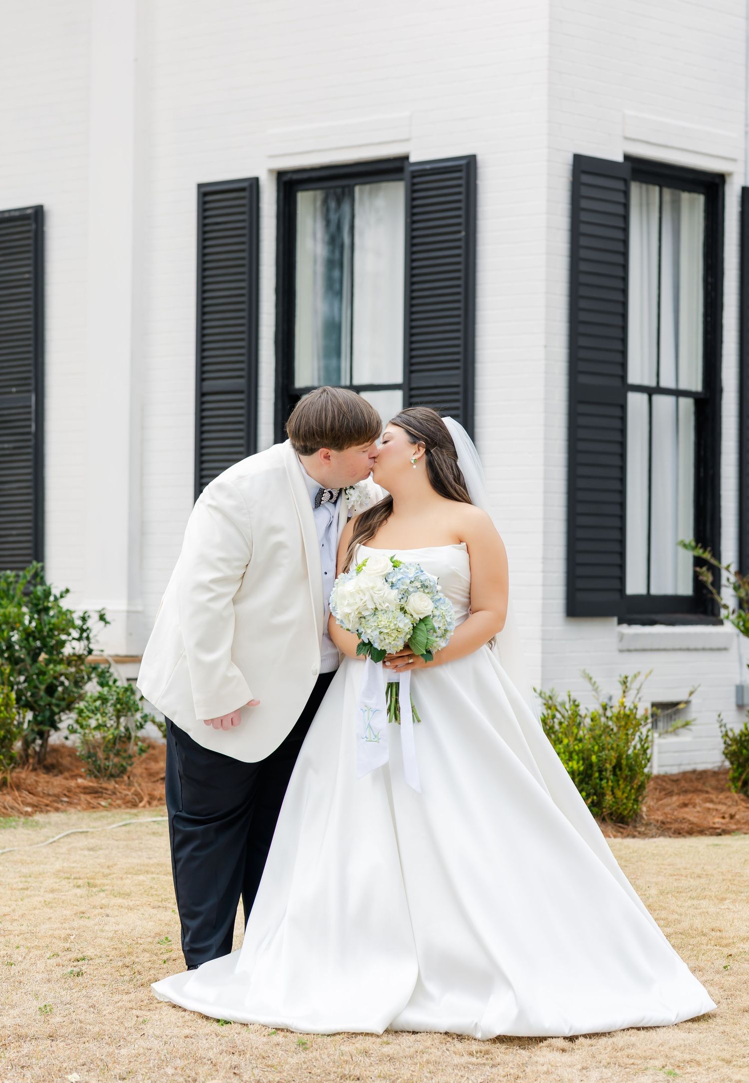 Bride and groom share a kiss outdoors in front of a white building with black shutters on their wedding day.