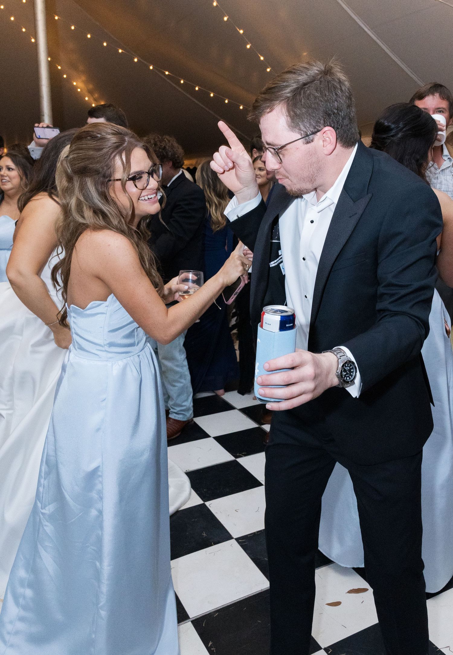 Wedding guests dance on a black and white checkered floor under a tent with string lights overhead.