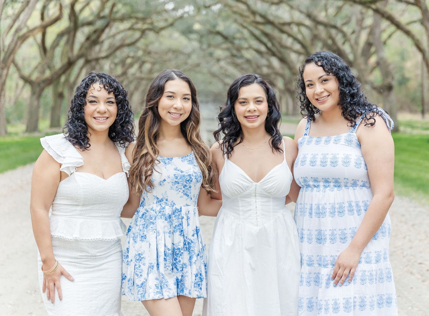 Four friends in white and blue dresses smiling together during an outdoor photoshoot.