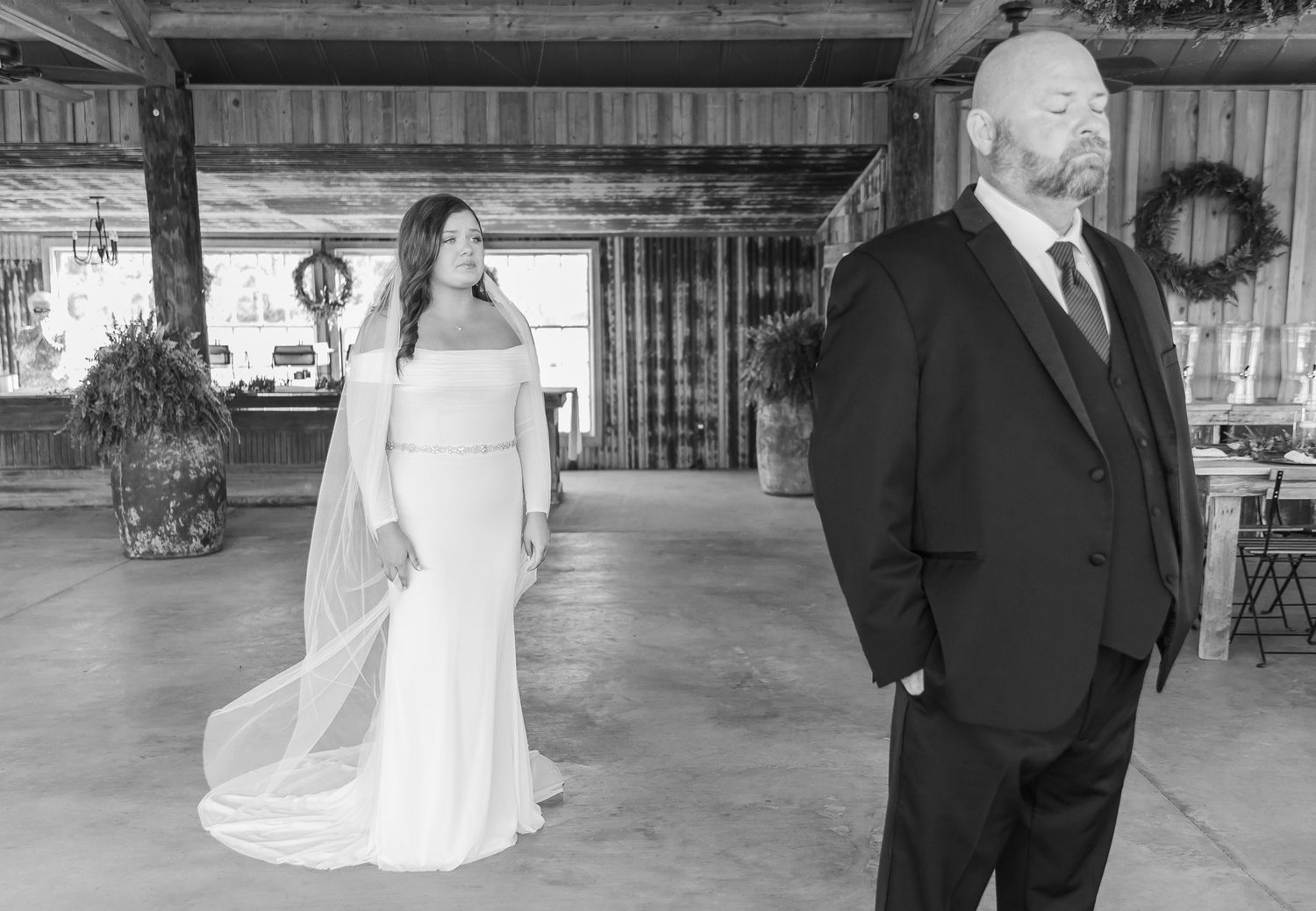 A black and white first look photo with a bride and suited figure standing in a rustic venue.