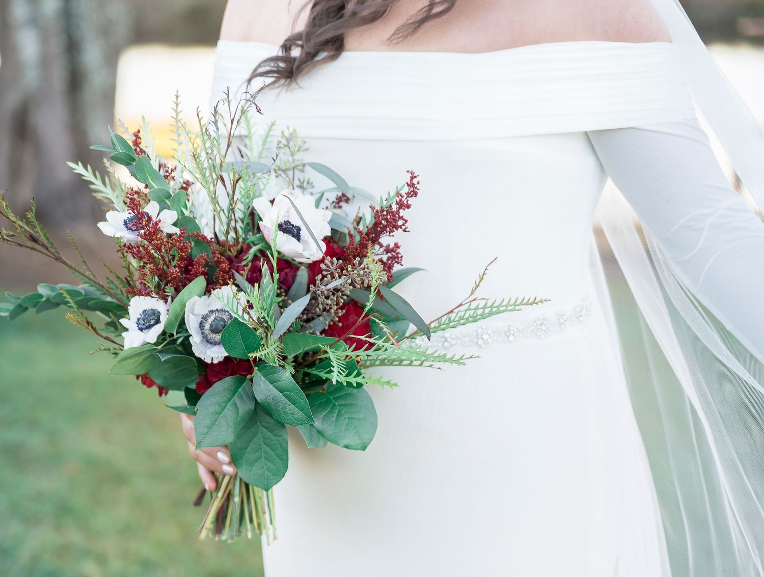 Winter bridal bouquet featuring white anemones, red berries, and greenery against a white dress.