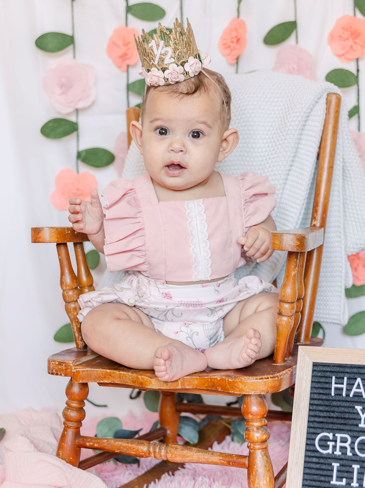 Baby in pink outfit and crown sits in wooden chair with milestone sign and floral background.