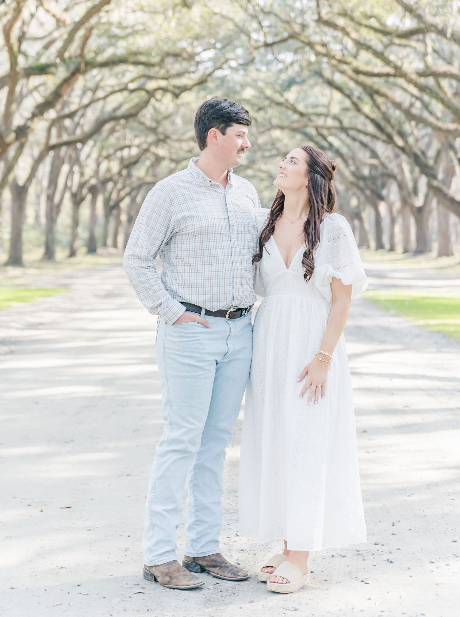 A couple in coordinating white and gray outfits stands on a scenic tree-lined path.