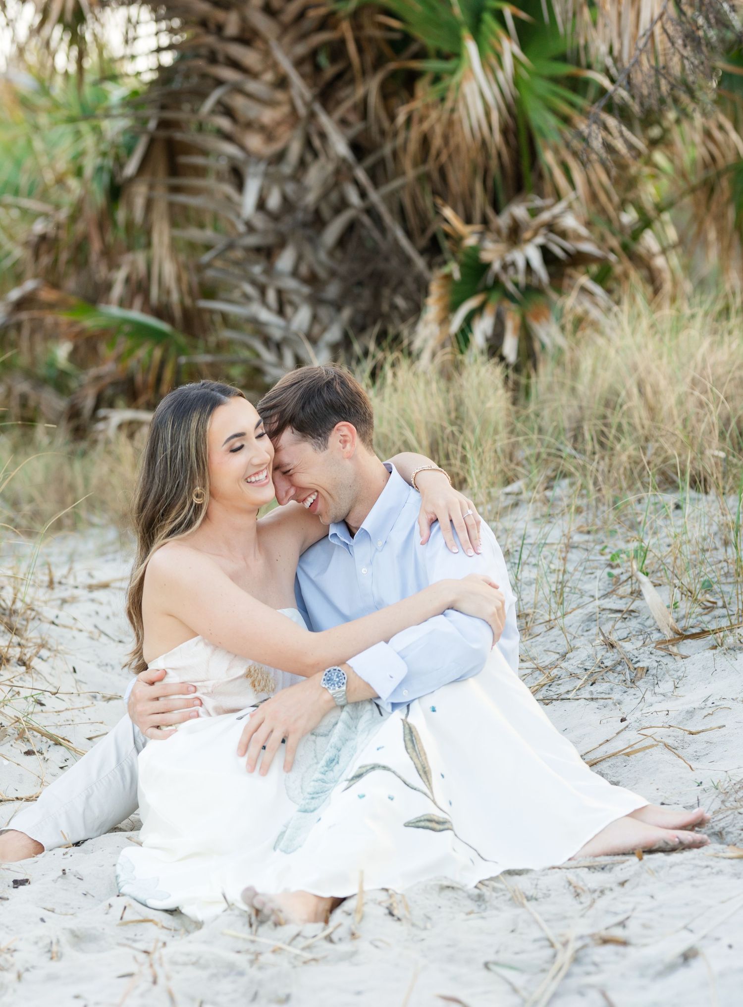 Happy couple embracing and laughing together on sandy beach surrounded by palm trees and coastal dunes.