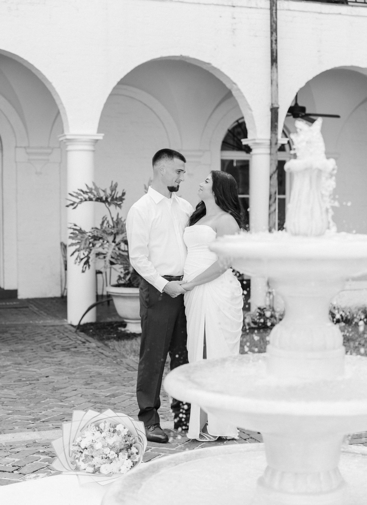 Black and white photo of a couple sharing a romantic moment near a fountain in an elegant courtyard with arched columns.