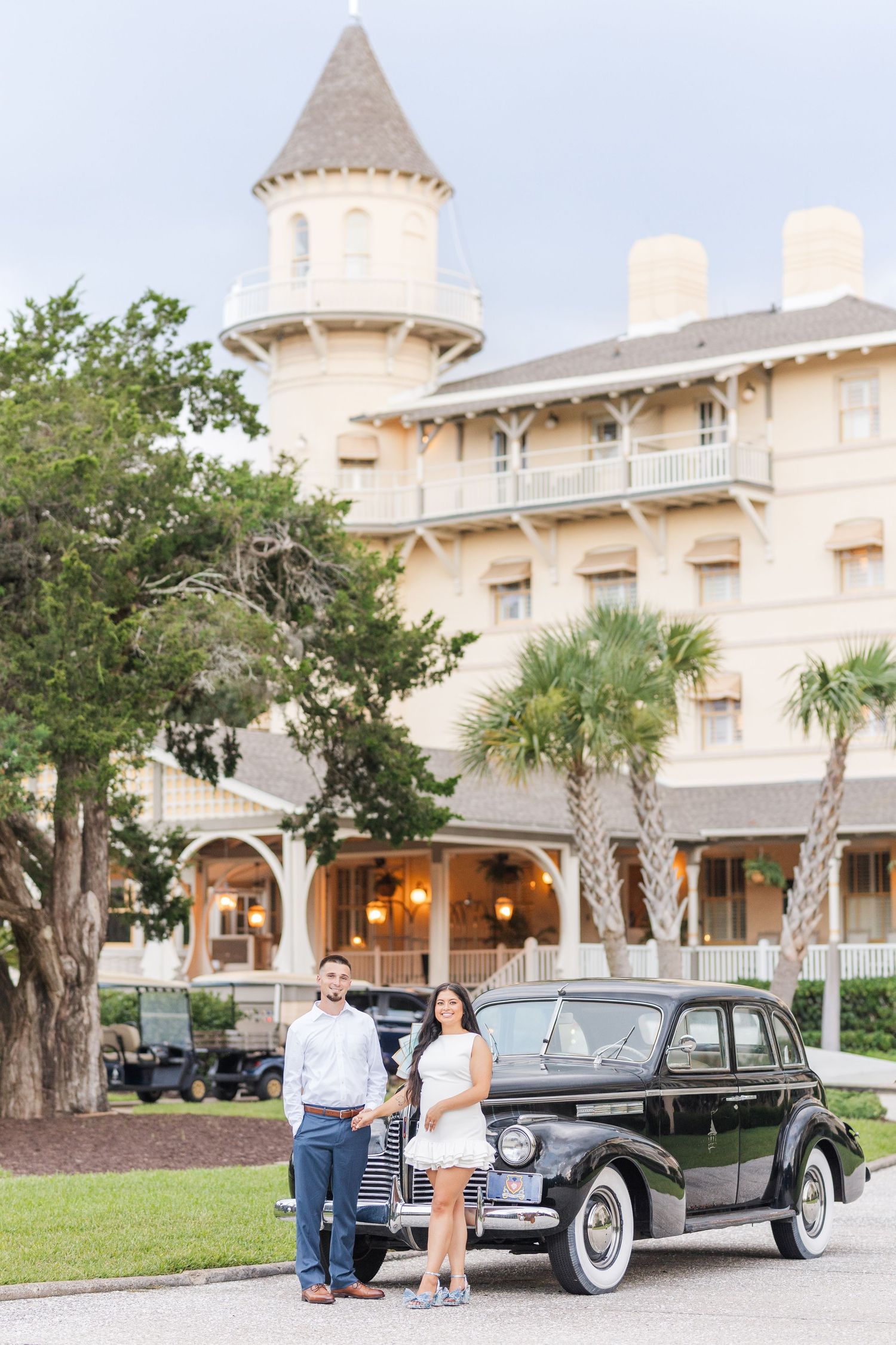 Engaged couple poses beside vintage black car in front of grand Victorian hotel with palm trees.