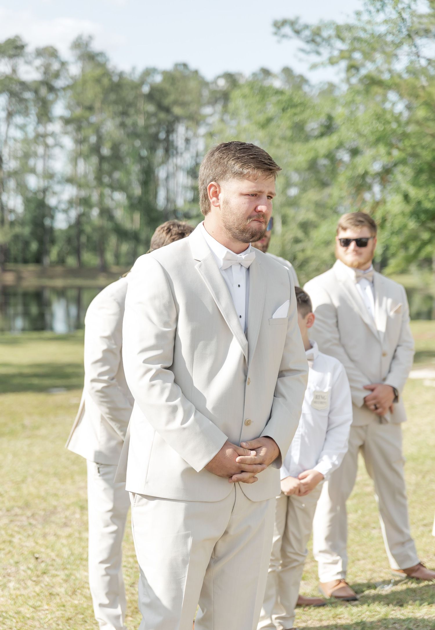 Groom in light colored suit stands nervously during outdoor wedding ceremony.