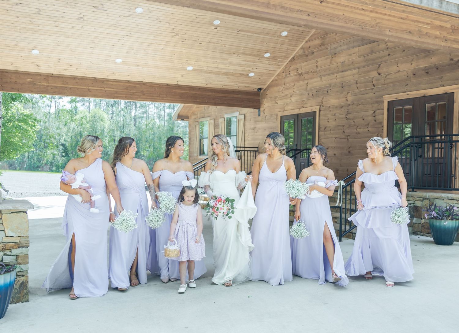 Wedding party in coordinated lavender dresses and white gown pose on wooden deck.