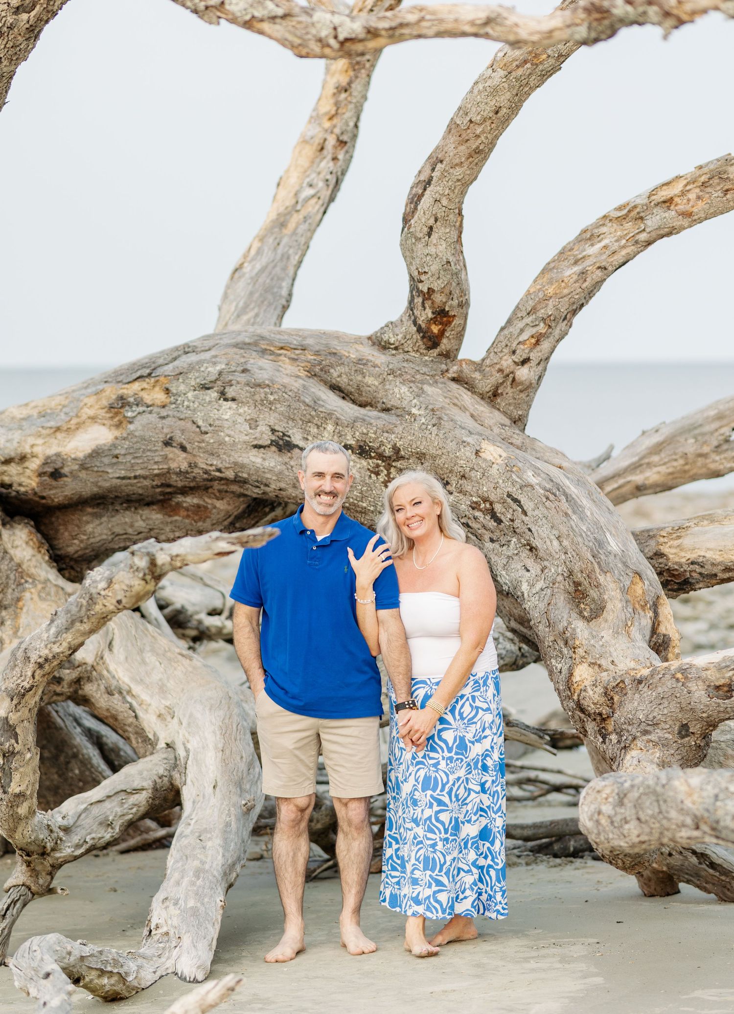 Couple poses together on a beach next to large driftwood during a sunny day with ocean background.