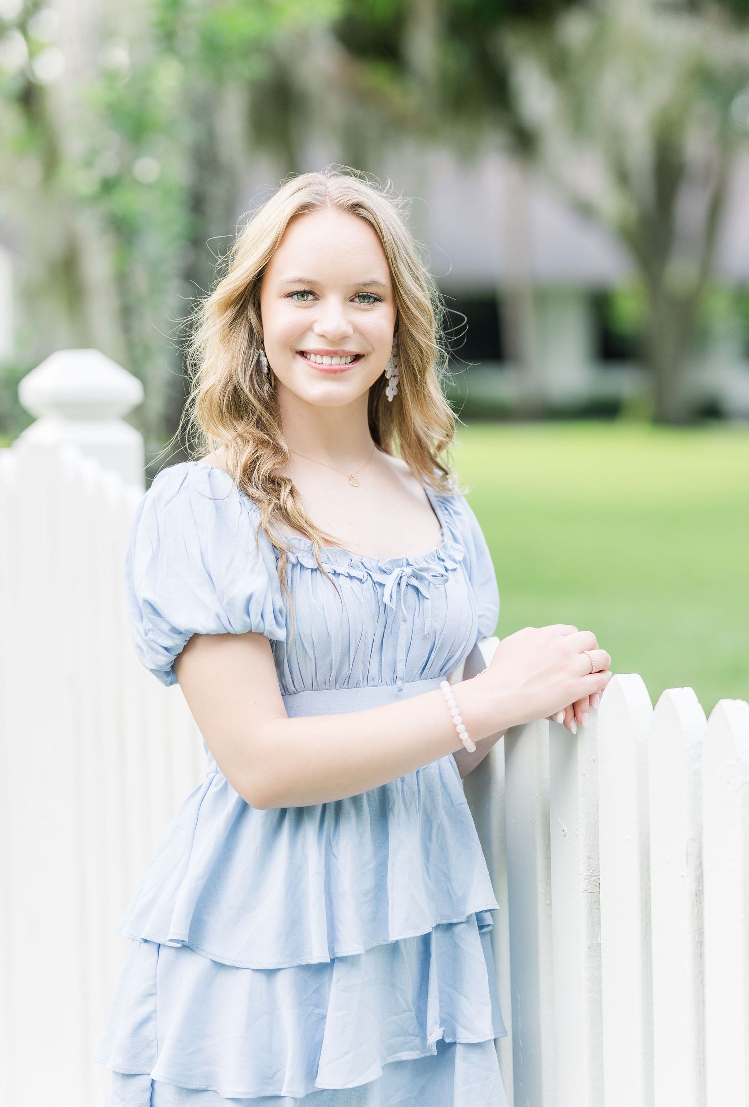 Woman in light blue dress poses by white picket fence in garden setting.