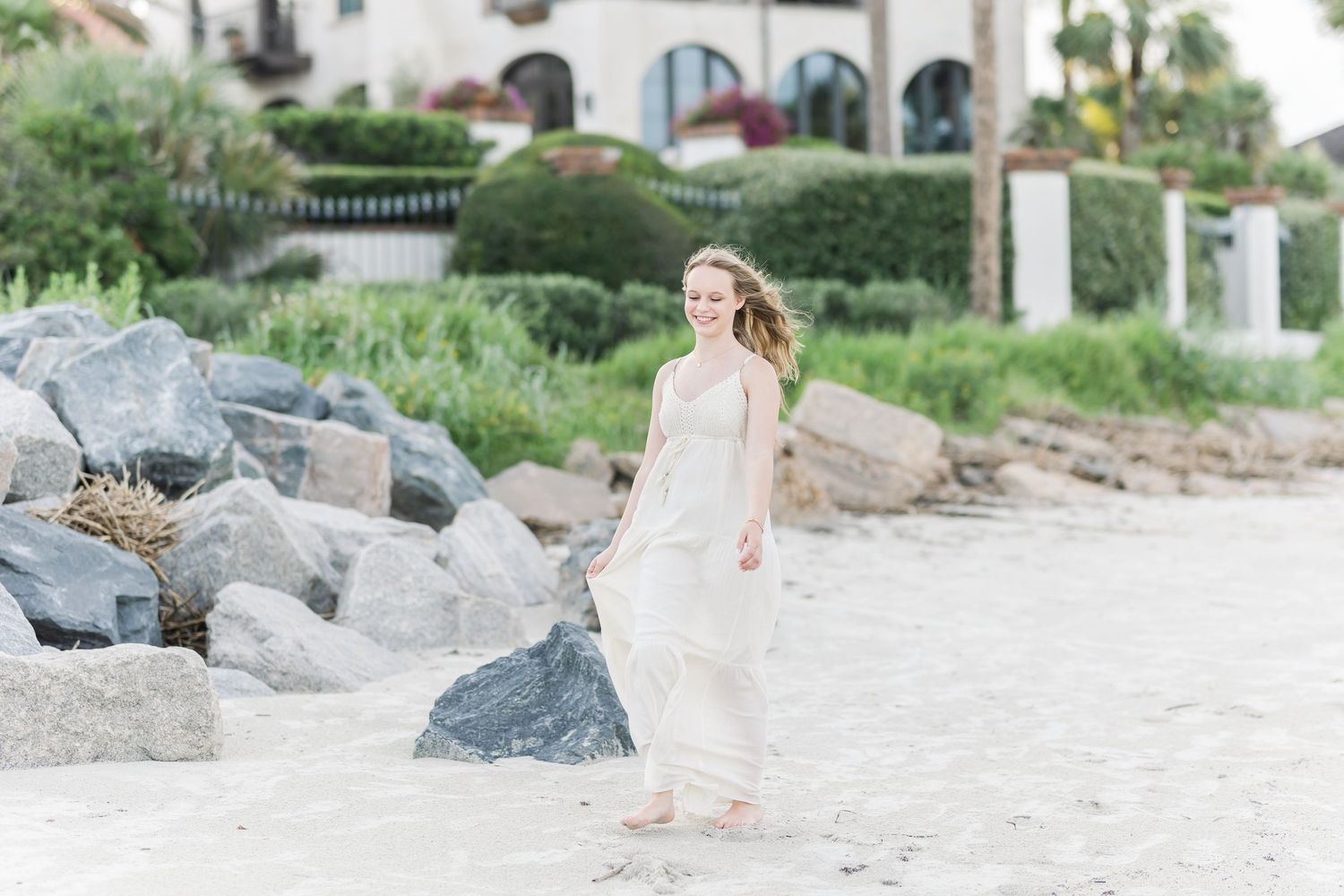 Person strolling along rocky coastal beach with Mediterranean style architecture in background.