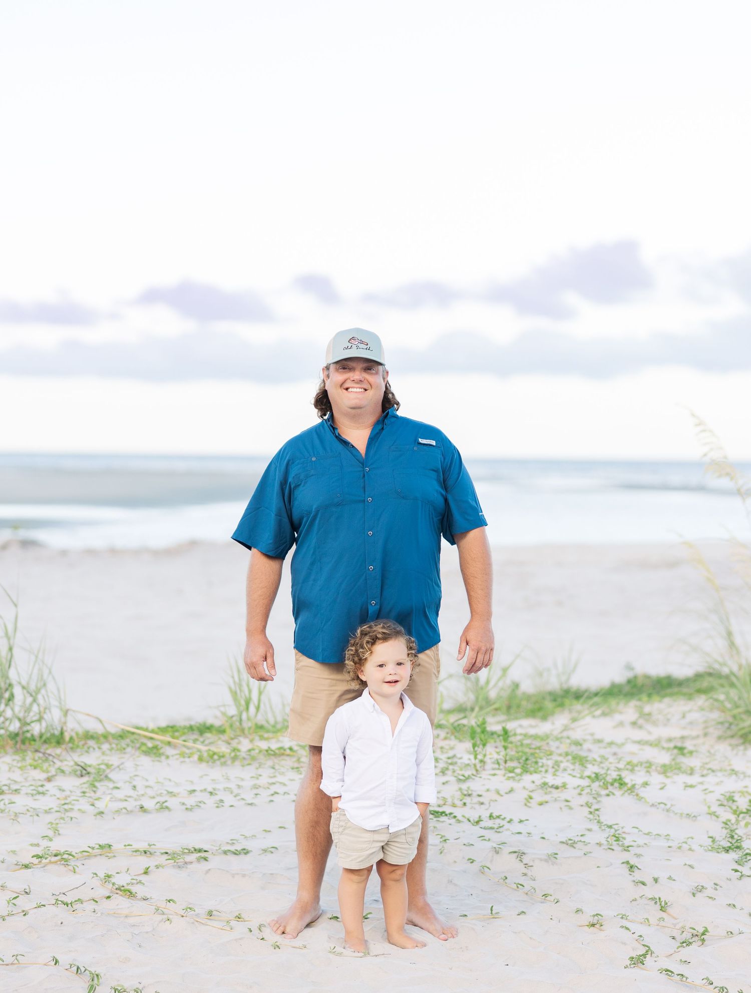 Adult in blue shirt and khaki shorts stands with small child on beach with ocean behind them.