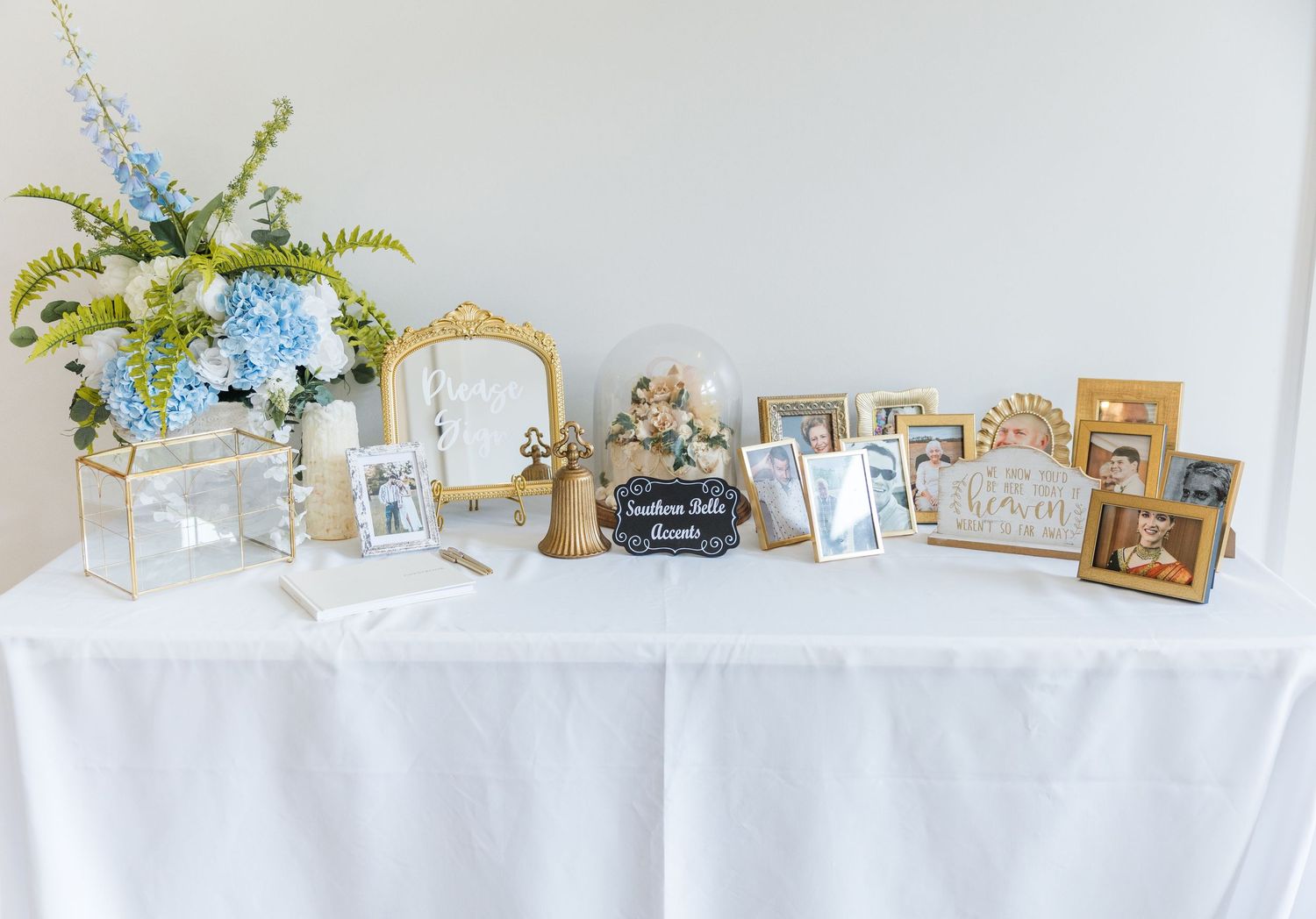 Elegant wedding gift table with blue hydrangeas, gold frames, photos, and a bell centerpiece on white linen.