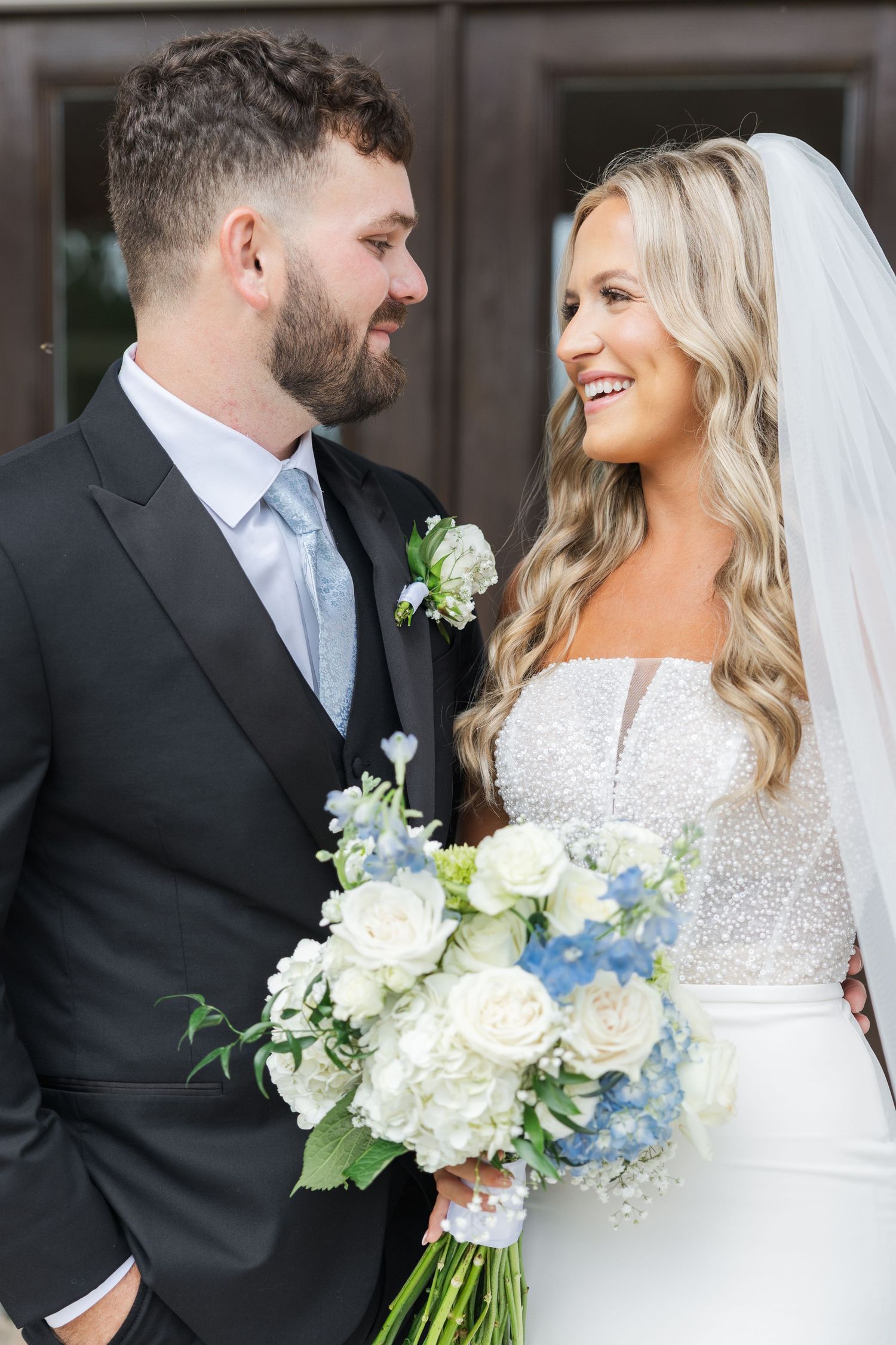 Smiling bride and groom share a romantic moment, bride holding a blue and white floral bouquet in elegant wedding attire.