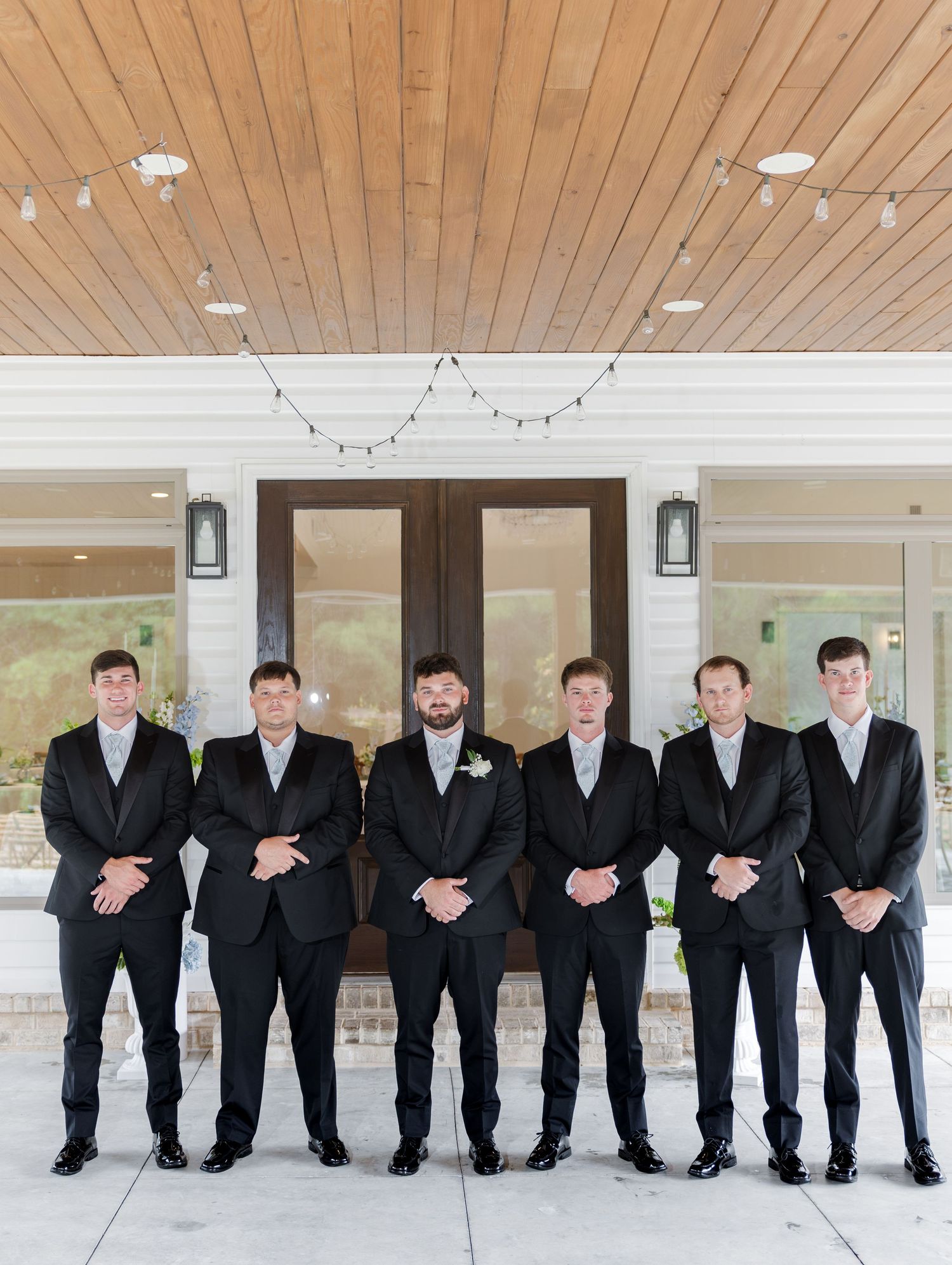 Groom and six groomsmen in black suits lined up in front of a wedding venue.